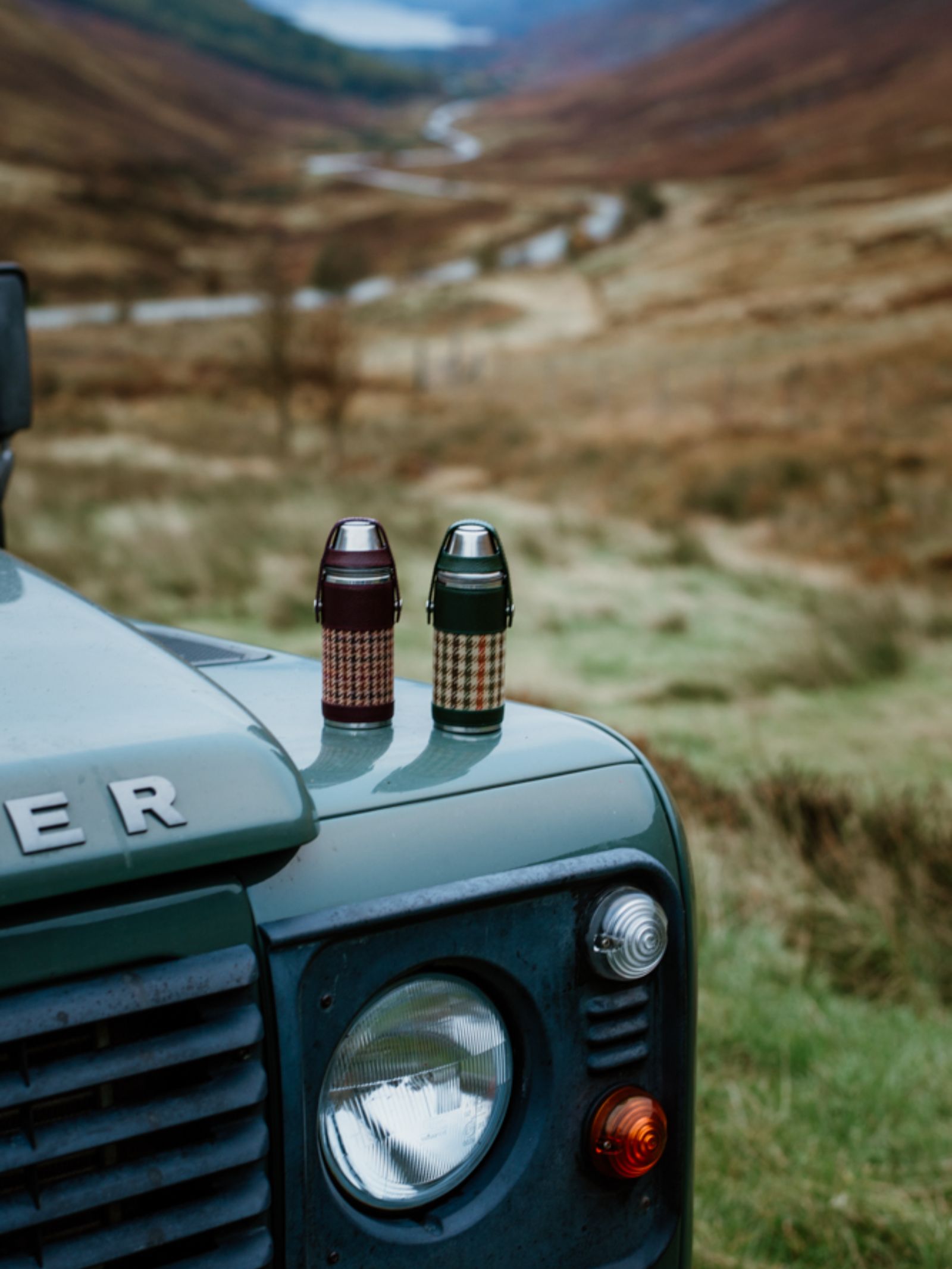 An 8oz Forest Green Hunter Flask by Campbell's of Beauly x Ettinger sits on a green Land Rover's hood, parked in grassy hills with a winding river below and clouds above—a scene that highlights British craftsmanship.