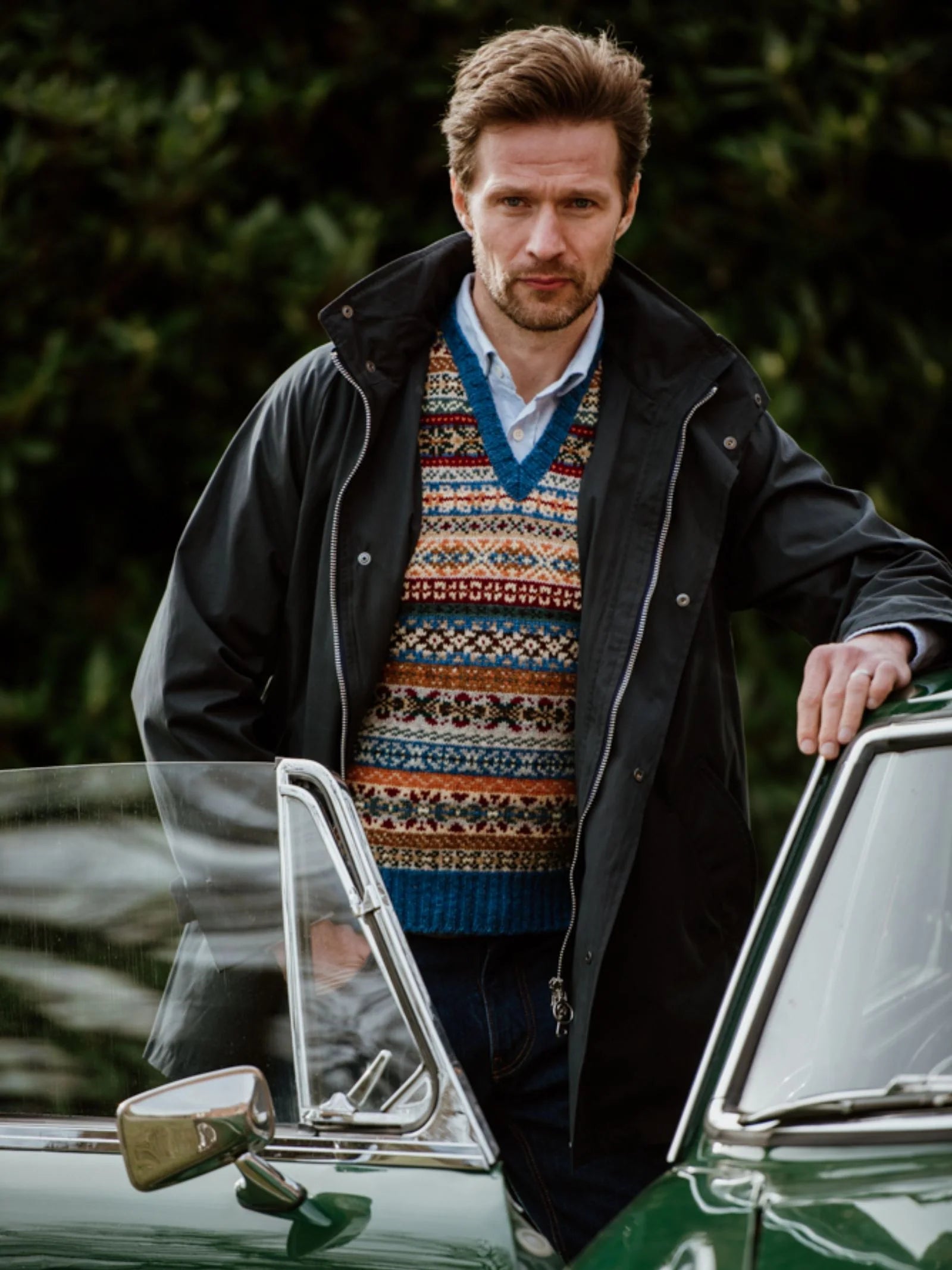 A man wearing the Campbell’s of Beauly Fairisle Slipover—crafted from soft Shetland wool—paired with a blue shirt and dark jacket, stands beside a green vintage car with its door open, set against a backdrop of greenery.