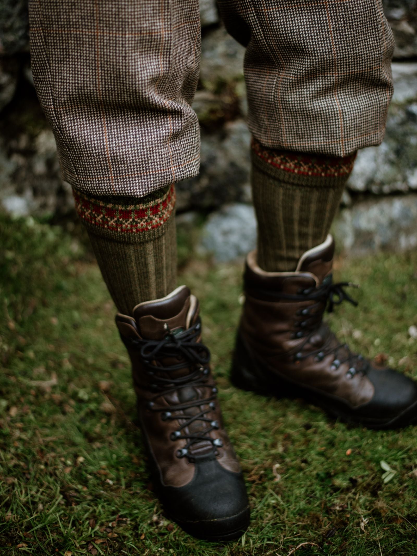 A person stands on grass wearing brown hiking boots, brown plaid knickerbocker trousers, and Campbells of Beauly Fairisle Shooting Socks in a merino-acrylic blend with red accents; only their legs appear before a blurred stone wall.