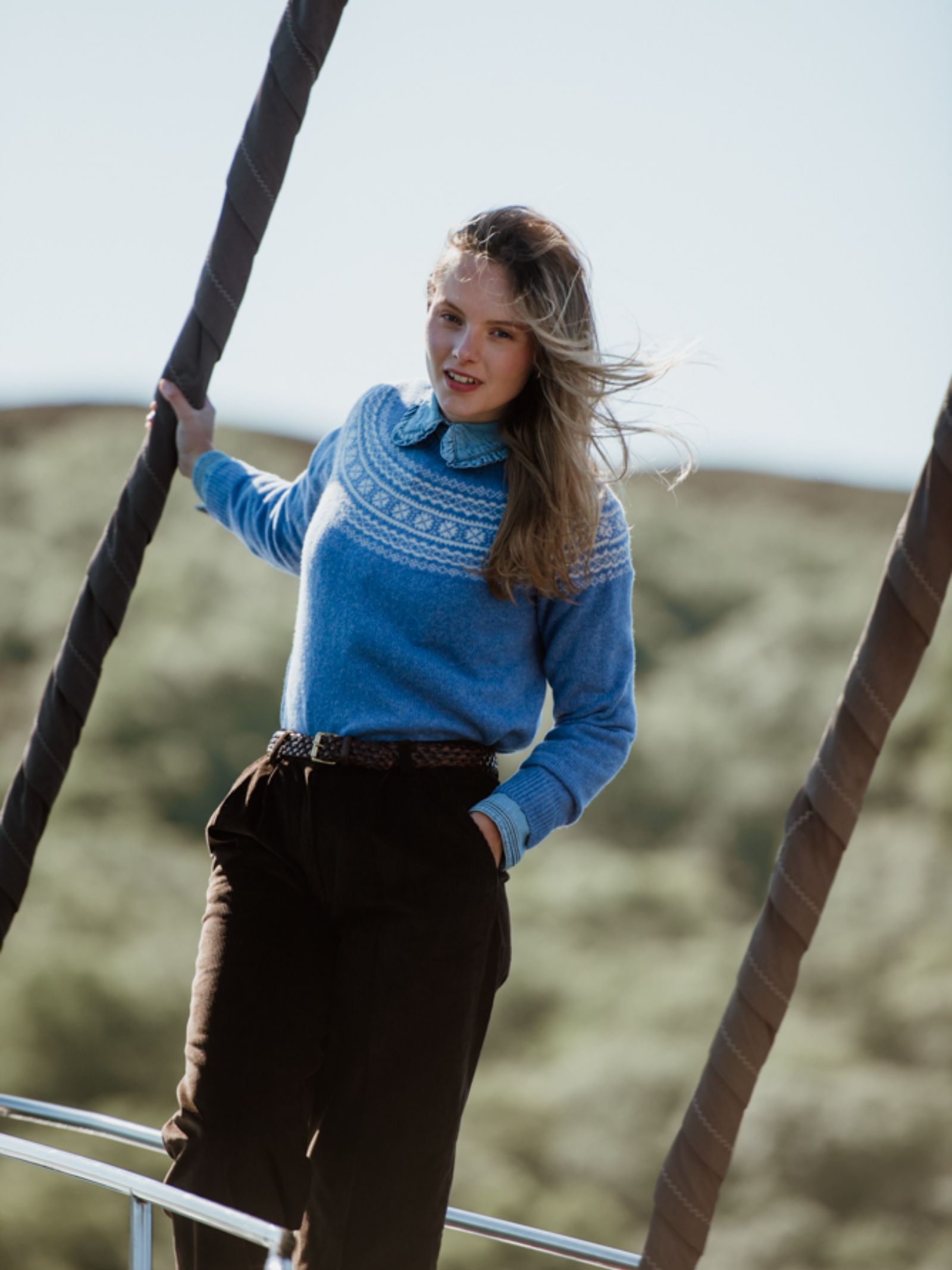 A woman stands outdoors holding two ropes, wearing Campbells of Beaulys Two-Colour Fairisle Yoke Jumper made from Superfine Geelong lambswool over a collared shirt, with loose blonde hair and blurred green hills behind her.