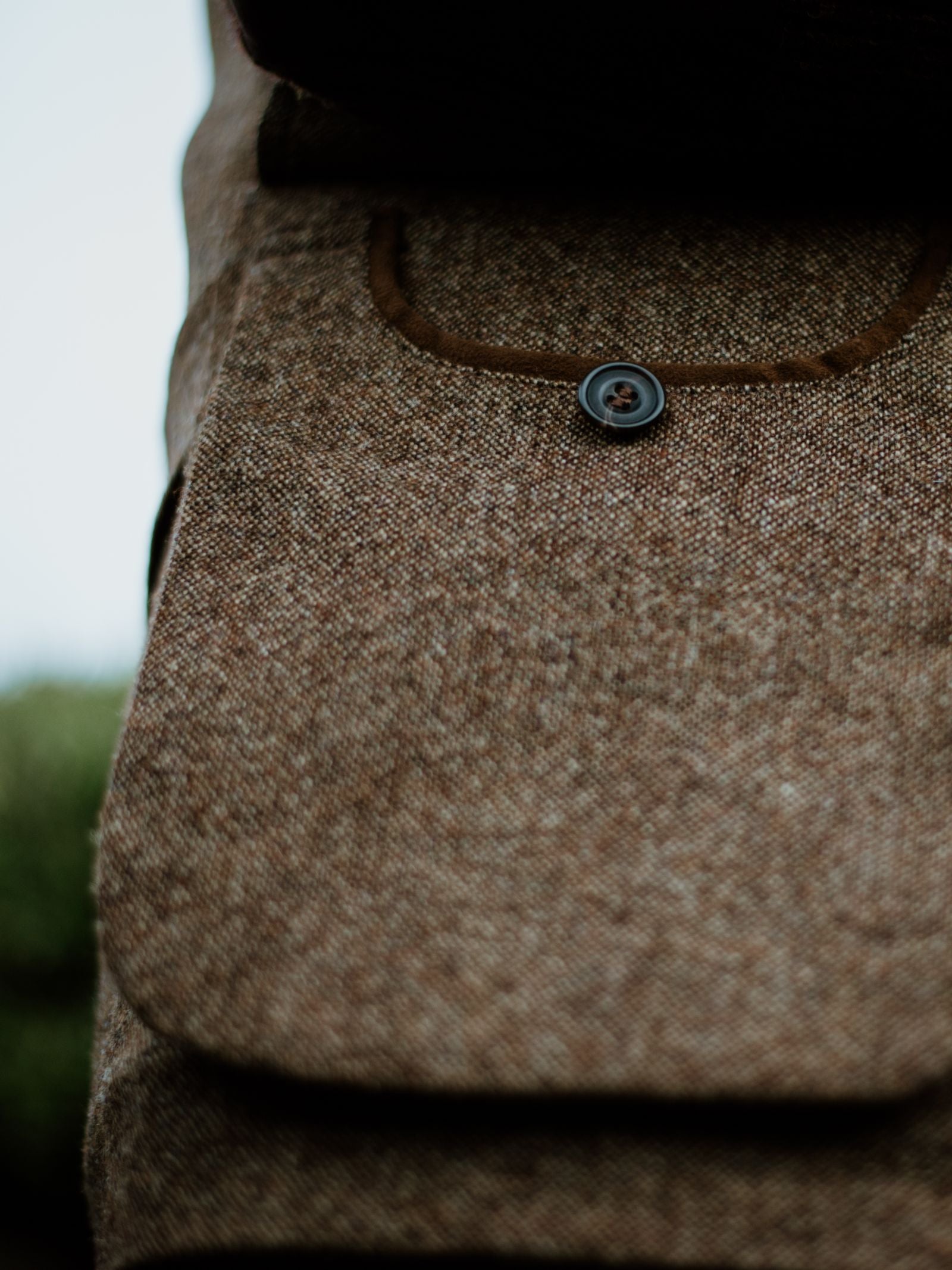 Close-up of the Field Nehru Gilet by Campbells of Beauly shows its textured brown tweed pocket with flap and black button, fine woven detail, and British moleskin accents, set against a blurred green outdoor background.