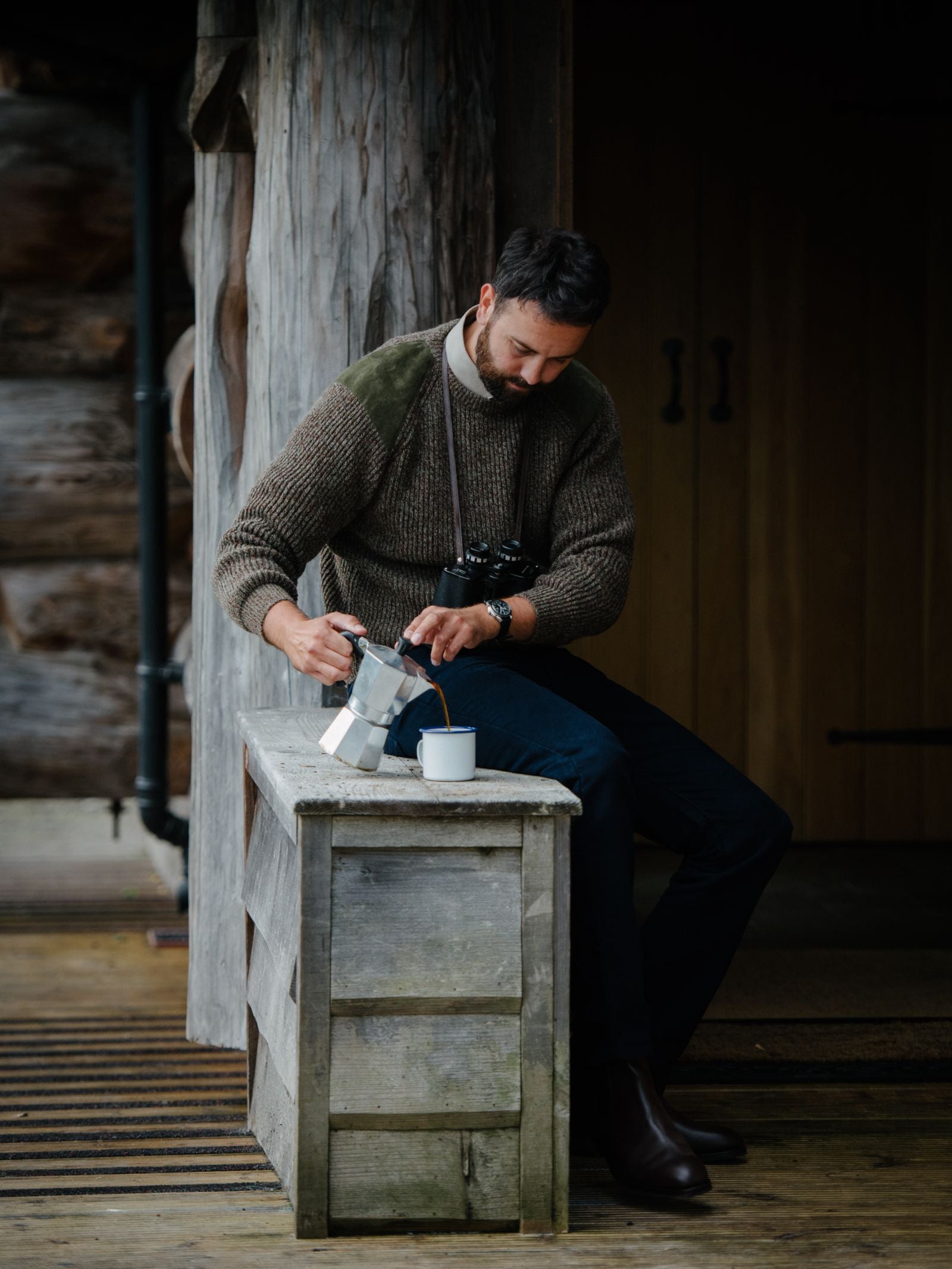 A bearded man sits outside on a rustic bench, pouring coffee from a moka pot into a mug. He wears the Campbells of Beauly Scotia Suede Jumper, dark pants, and boots. A camera hangs around his neck near a wooden cabin and large door.