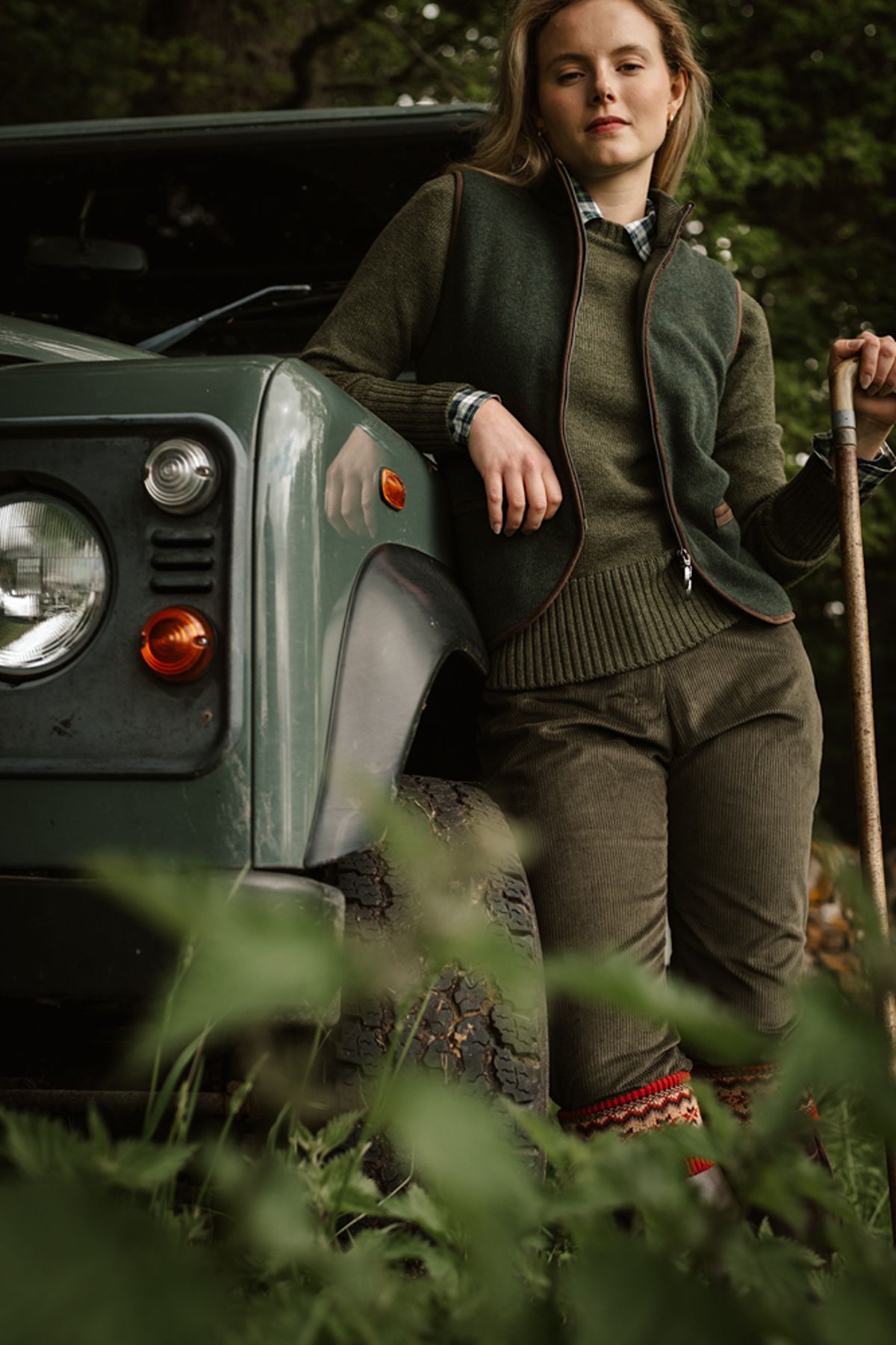 A woman in a green jumper vest and trousers leans against a green vintage off-road vehicle whilst holding a walking stick.