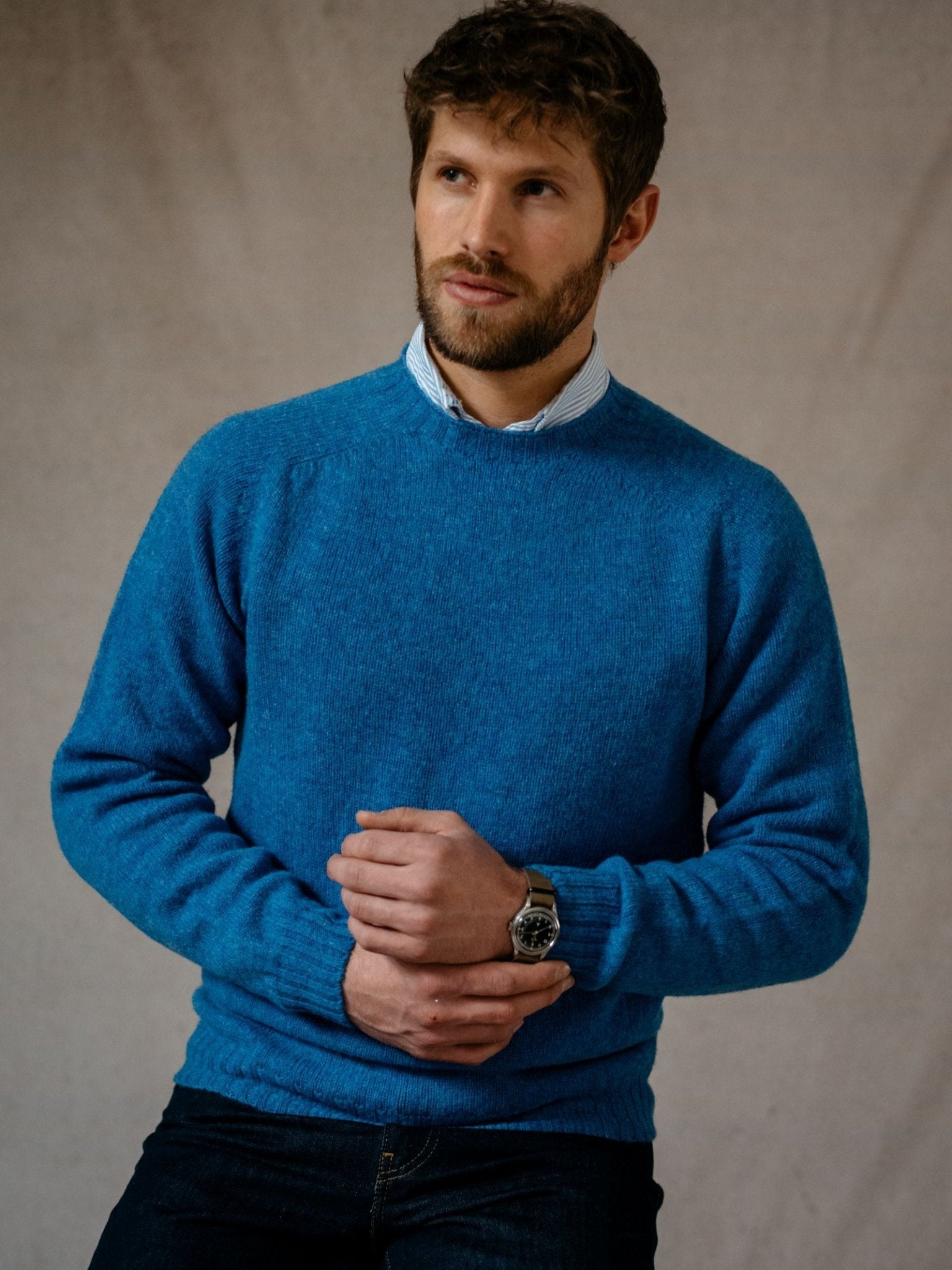A man with short brown hair and a beard wears a bright blue Campbells of Beauly Shetland Jumper over a collared shirt and dark jeans, standing against a neutral background with his hands clasped in front of him.