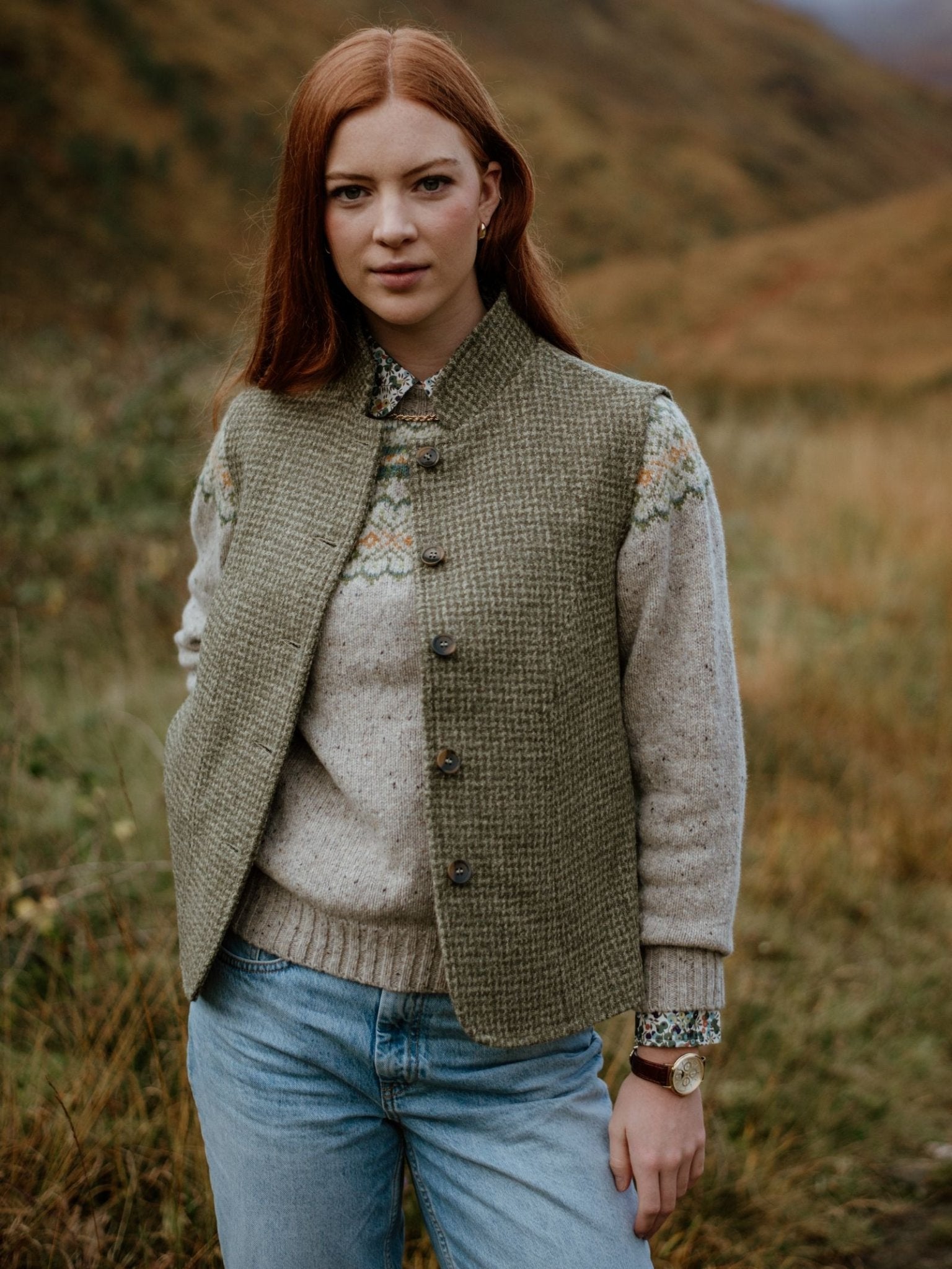A woman with long red hair stands outdoors in a grassy, hilly landscape. She wears a knitted sweater, blue jeans, and the Campbells of Beauly Harris Tweed Houndstooth Gilet, looking calmly at the camera.
