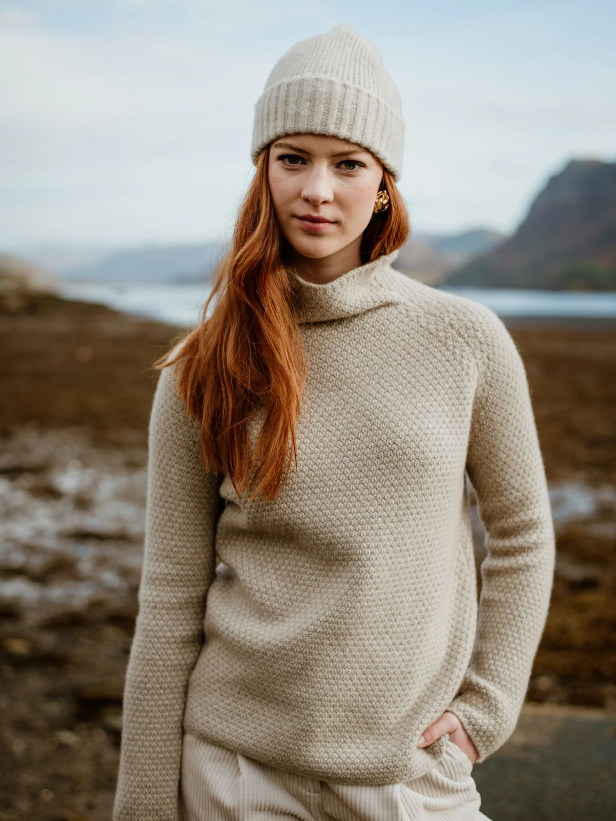 A woman with long red hair, wearing a Scottish knit sweater and a Campbell’s of Beauly Lambswool Hat Cream, stands outdoors by a rocky shore with mountains in the background.
