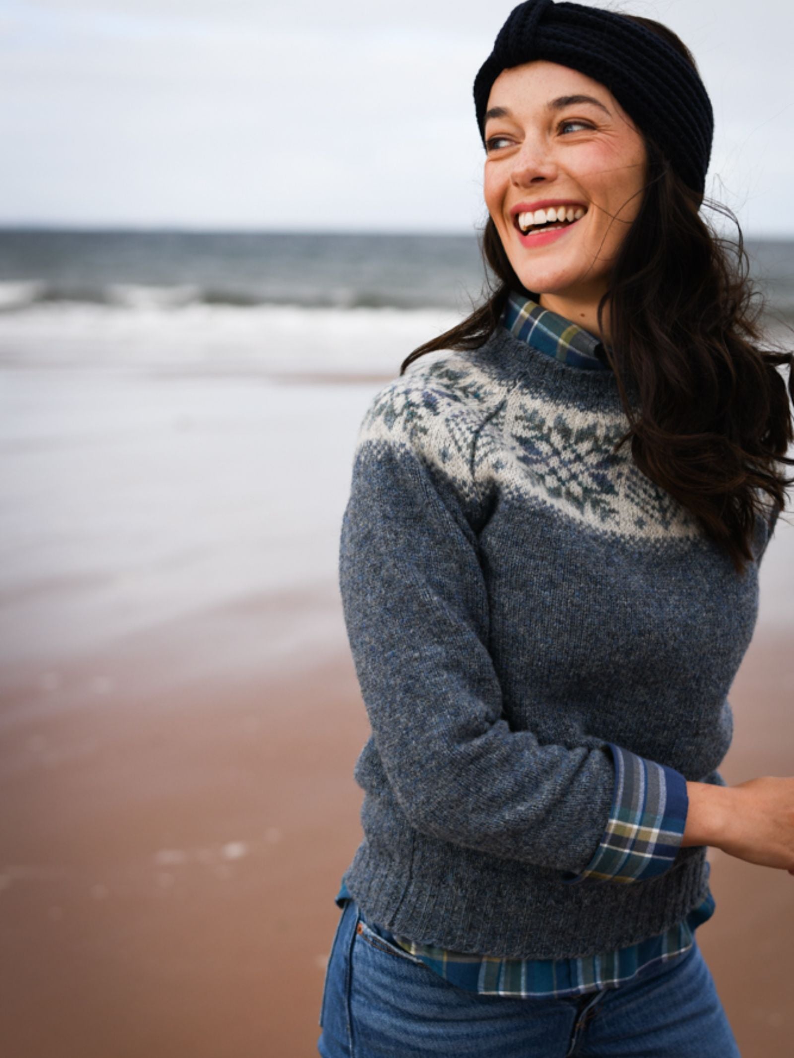 A woman wears the Campbell's of Beauly Shetland Wool Snowflake Crew Jumper with a plaid shirt, jeans, and a black headband, smiling as she stands on a sandy beach with the ocean behind her.