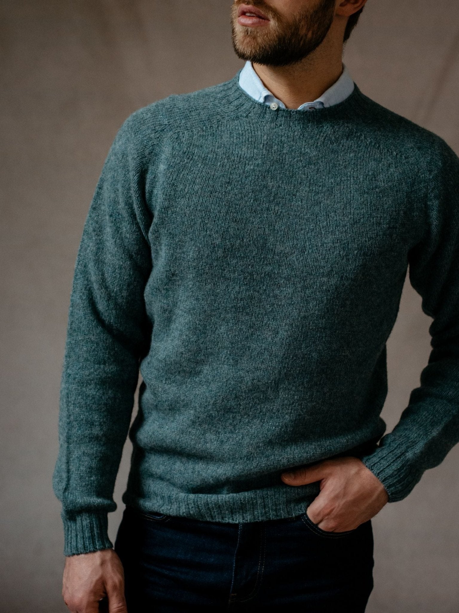A man wears a light blue collared shirt under a greyish-green Campbells of Beauly Shetland Jumper made from lambswool yarns, paired with dark jeans. He stands against a plain background, one hand in his pocket, face partially out of frame.