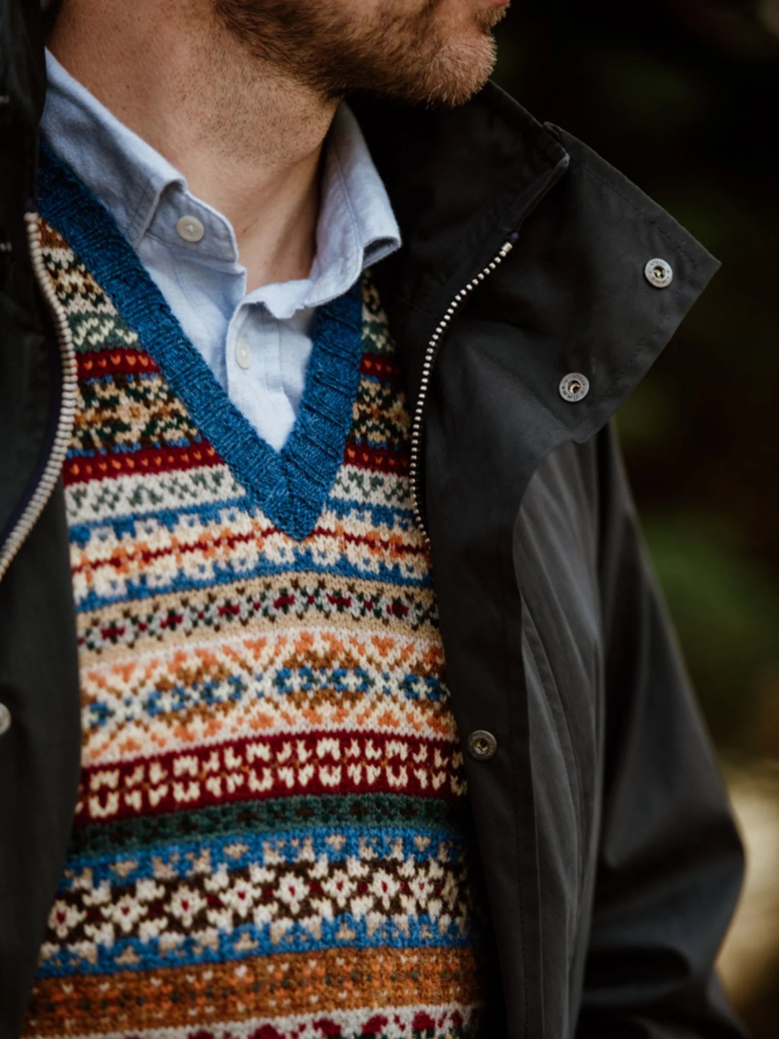 A man, slightly turned away from the camera, wears a blue collared shirt layered beneath Campbell’s of Beauly Fairisle Slipover—crafted from Shetland wool with vibrant patterns and finished with a blue trim—paired with a dark zip-up jacket.
