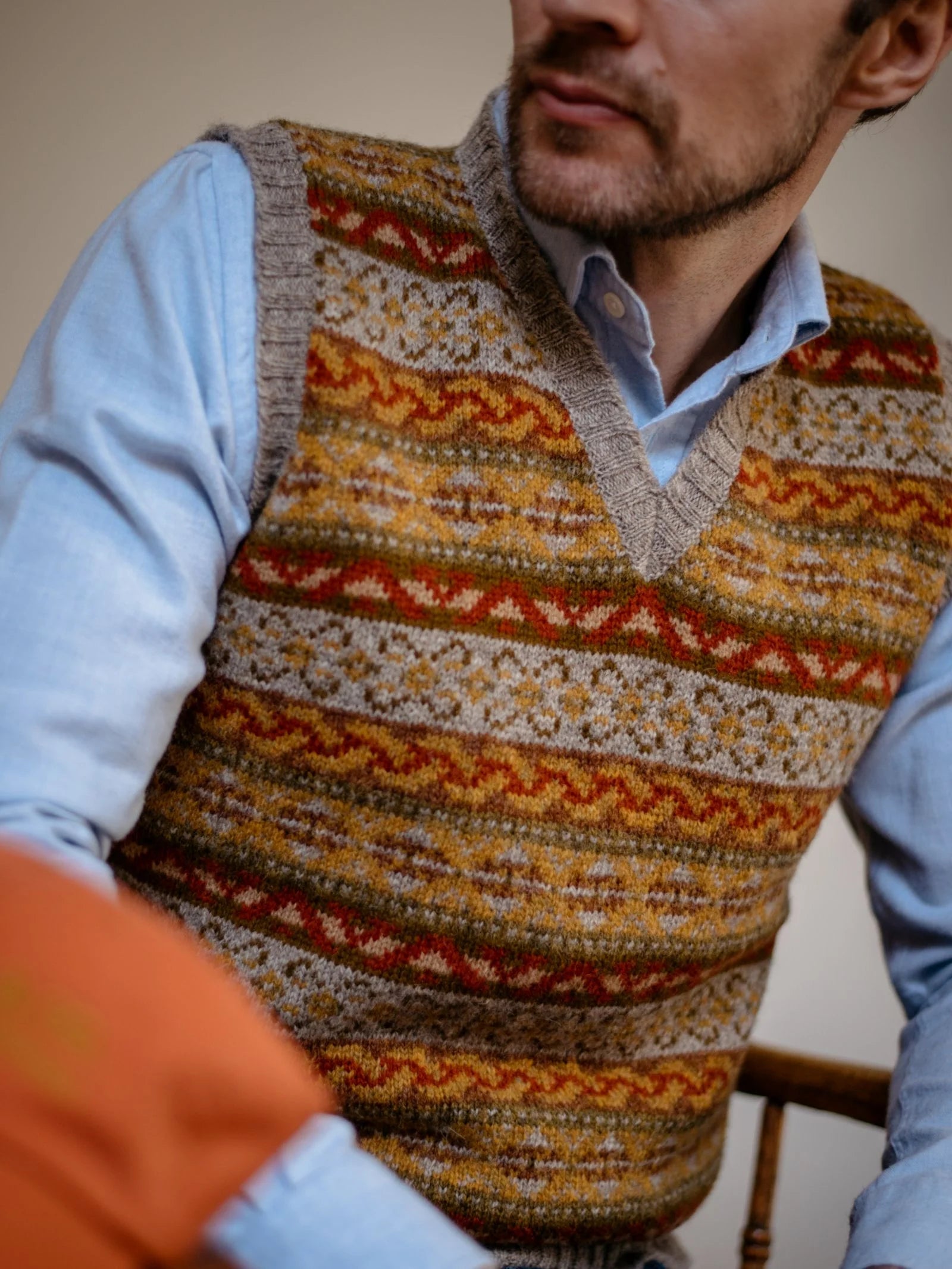 A man in a light blue shirt wears the Campbell’s of Beauly Fairisle Slipover, a knit sweater vest featuring colorful patterns in warm tones, as he sits on a chair, pictured from shoulders to waist.