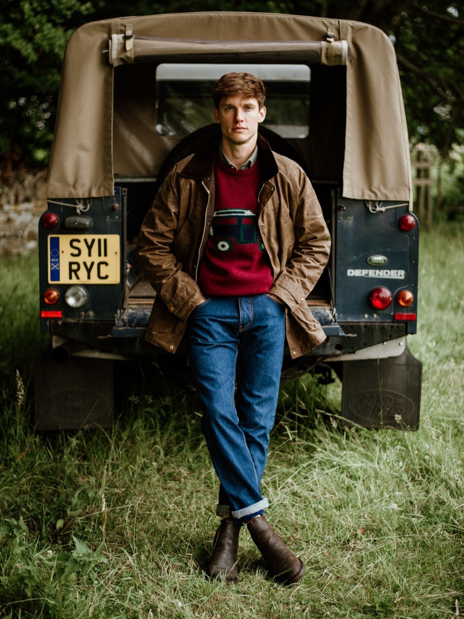 A man stands in front of a green Land Rover Defender with a tan soft top, wearing a brown jacket, red car-design sweater, blue jeans, and Campbell's of Beauly Lowlander Boots—handmade in England—on grass with trees behind.