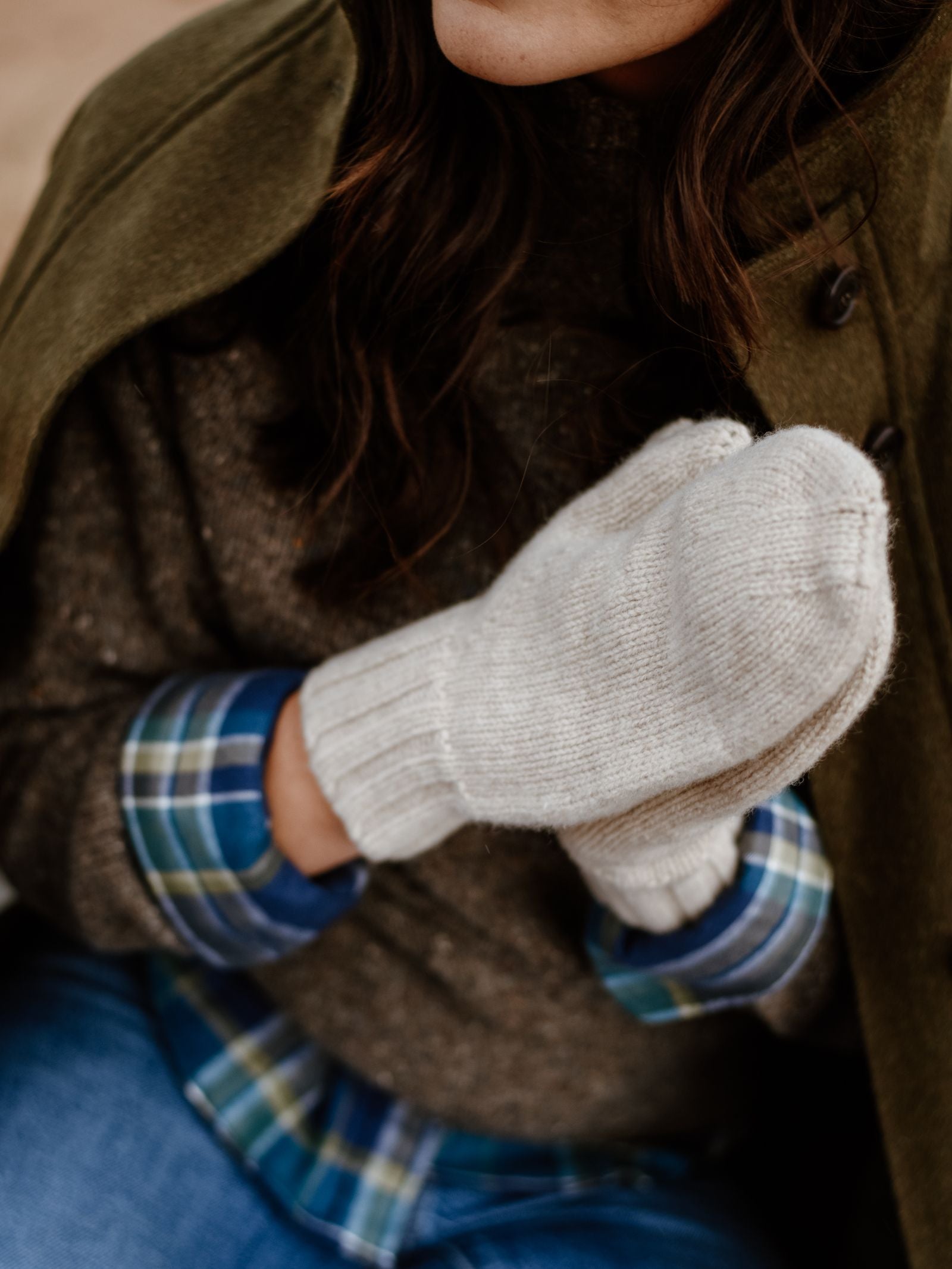 A person in a green coat, blue plaid shirt, and Campbell's of Beauly Chunky Mittens sits with hands together; mittens are gray lambswool, partially visible from the shoulders down.