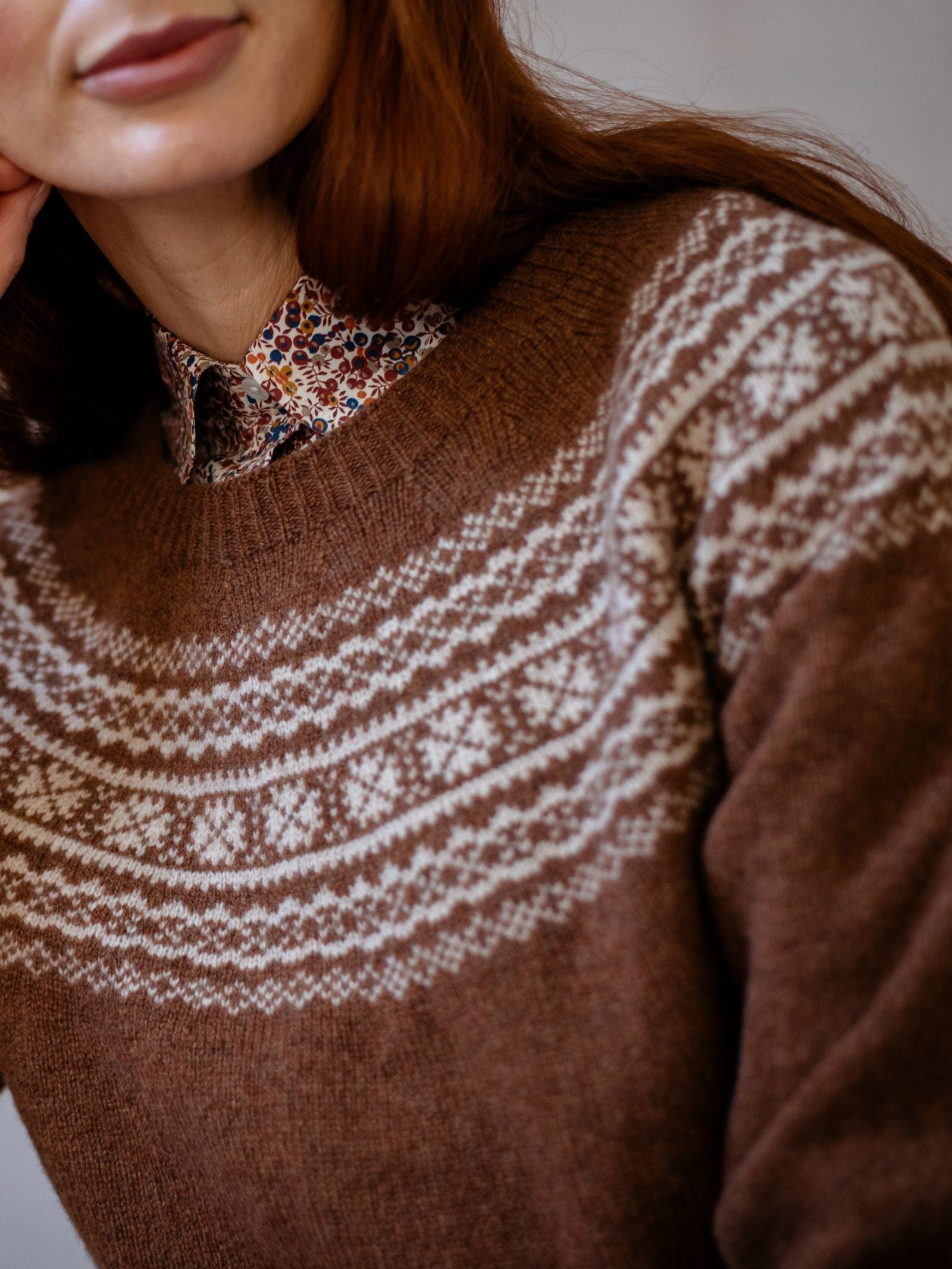 A woman with long reddish hair wears Campbells of Beaulys Two-Colour Fairisle Yoke Jumper, featuring a white Nordic pattern. A small, colourful floral print shirt collar is visible under the sweater. Only her lower face and upper torso are shown.