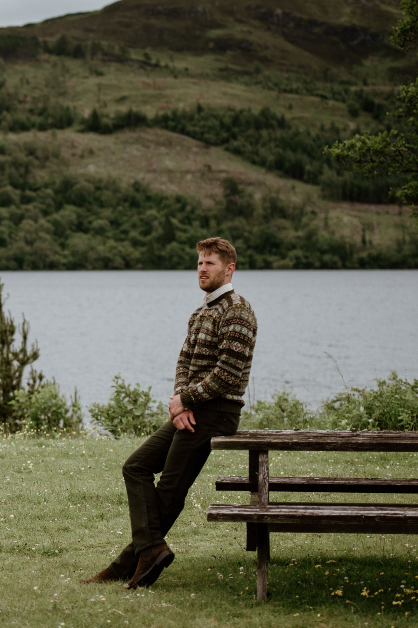 A man in earth-toned knitwear and Campbells of Beauly Moleskin Jeans with a classic 5-pocket design stands by a wooden picnic table near a lake, with green hills and trees under an overcast sky.
