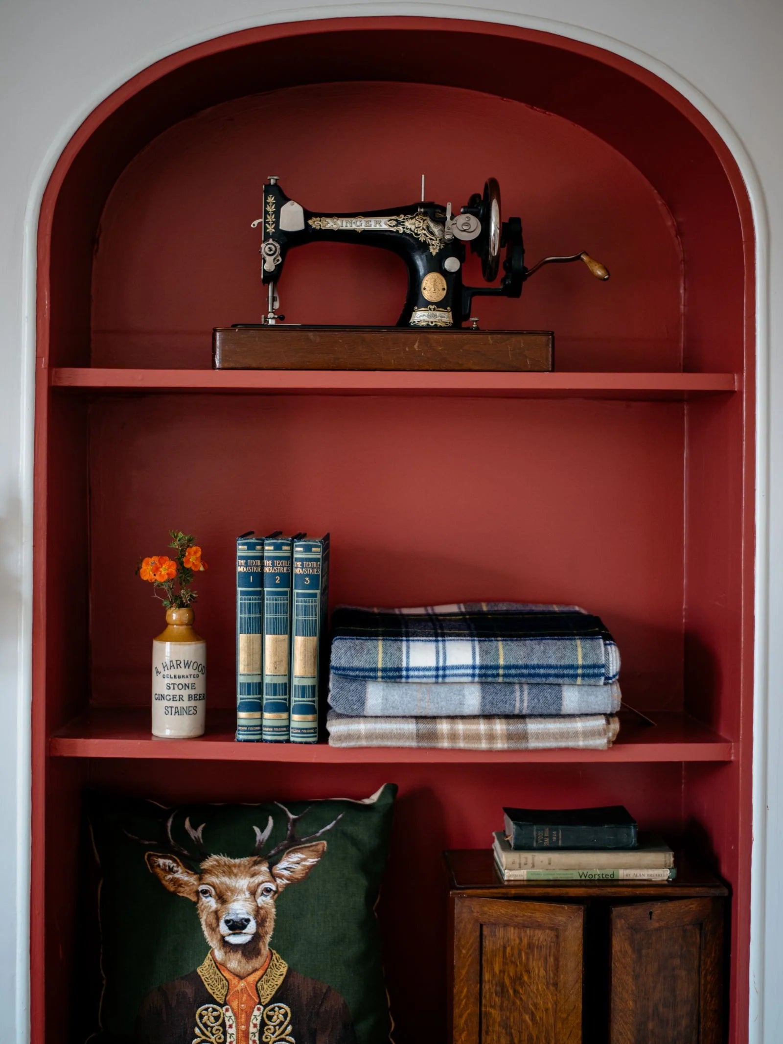 A vintage sewing machine sits on the top shelf of a red alcove bookshelf, with books, a soft blanket, the Campbell's of Beauly Lambswool Tartan Blanket, a small vase of flowers, and a pillow featuring a deer in a suit on the other shelves.