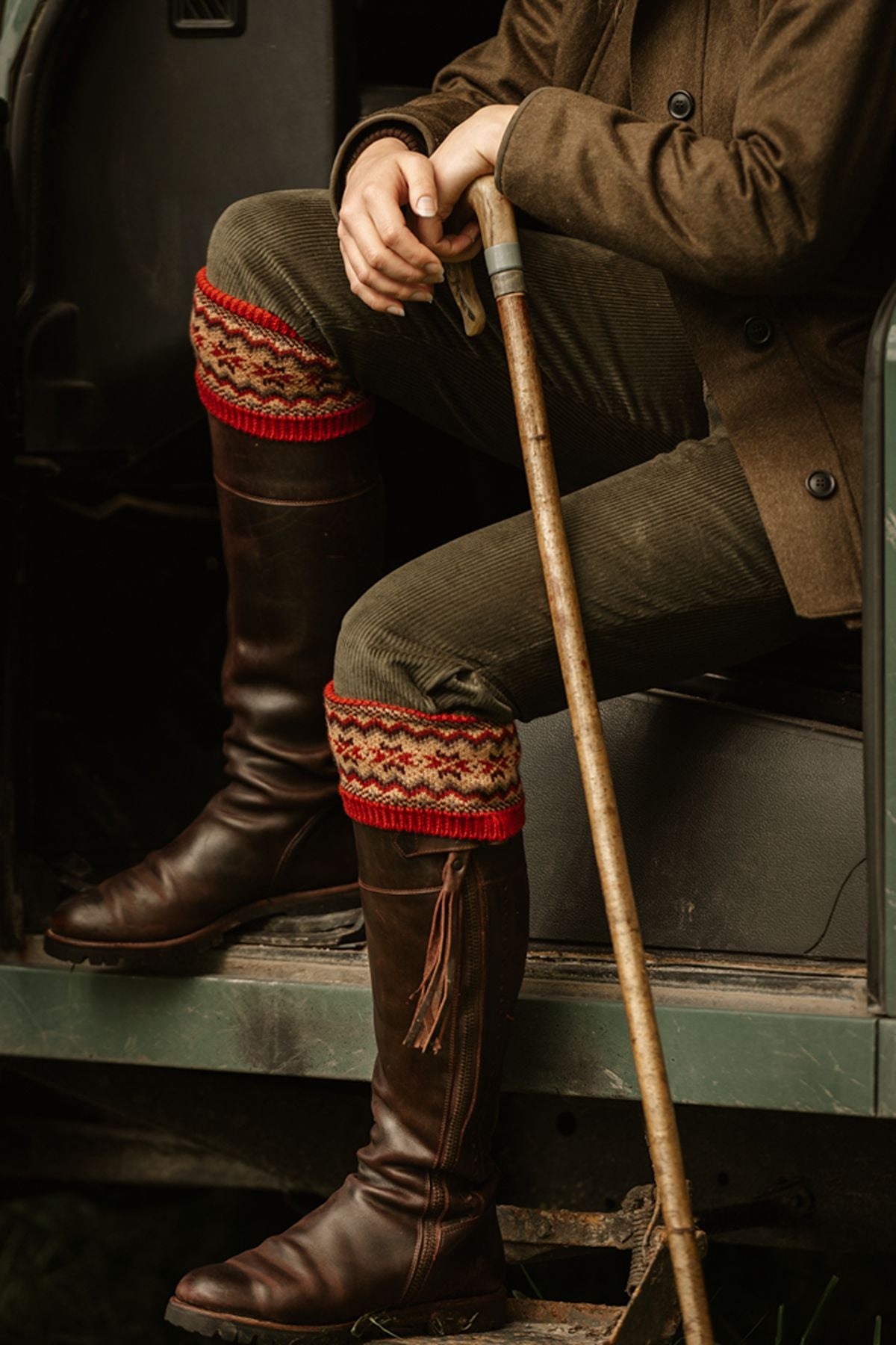 A person sitting in a vehicle wearing brown corduroy trousers, tall leather boots, and patterned red and tan boot cuffs, holding a wooden walking stick.