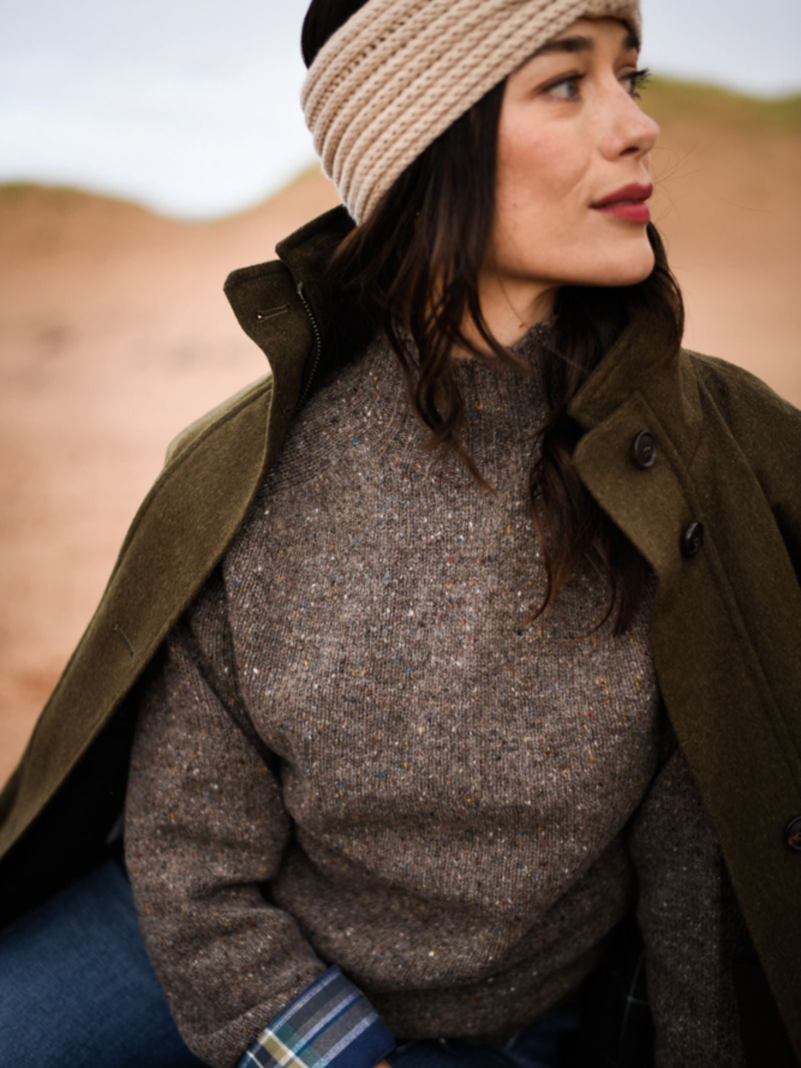 A woman in a beige knit headband, olive green coat, and a gray Campbell's of Beauly Donegal Lambswool Funnel Neck Jumper sits outdoors on sandy terrain, gazing calmly to the side.