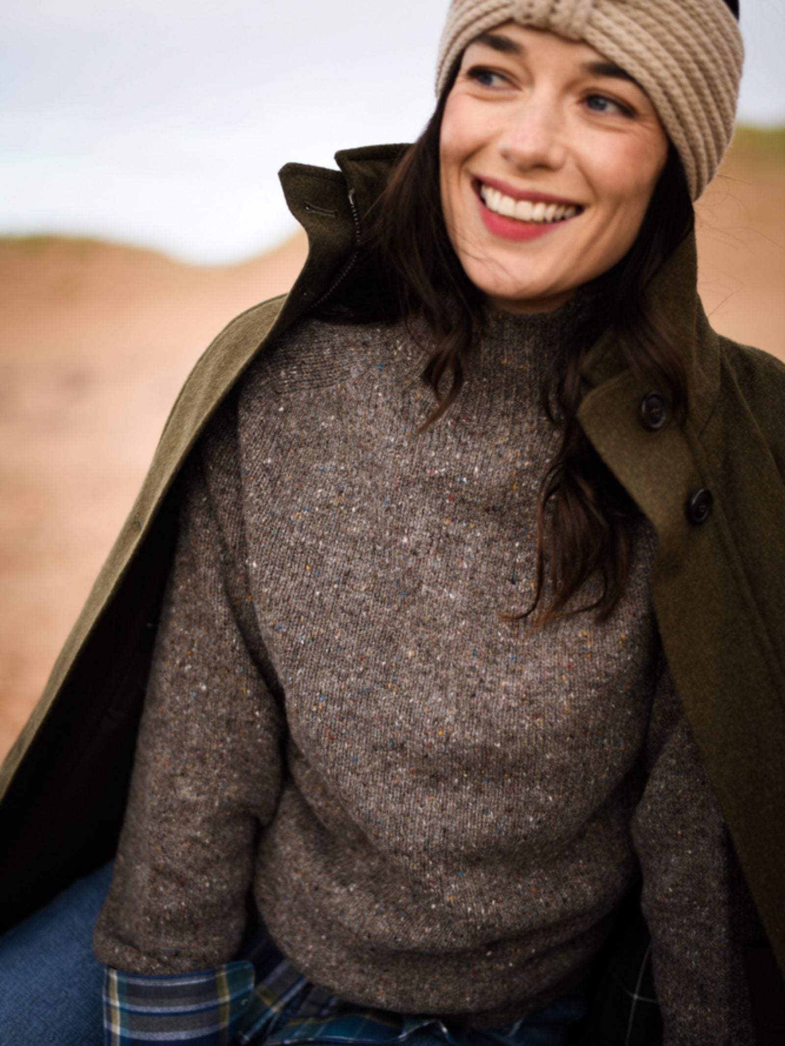 A woman smiles outdoors in a dark green coat, brown knit sweater, and plaid pants, finishing her look with the Campbell’s of Beauly Cashmere Ear Warmer—the perfect winter accessory against a rugged landscape.