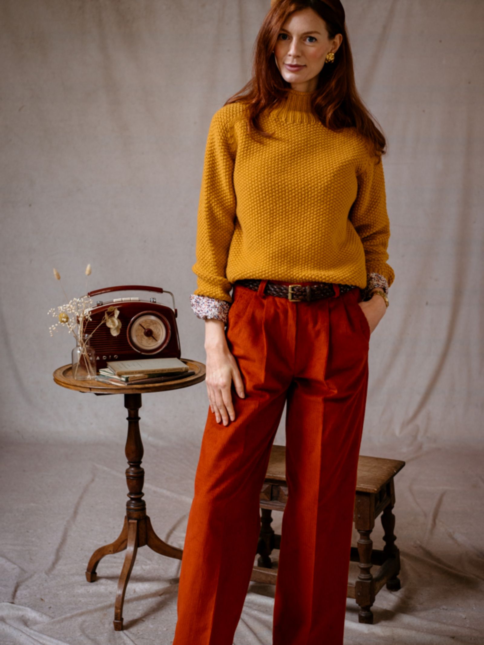 A woman with long red hair wears Wide Leg Cords by Campbells of Beauly and a mustard yellow sweater, standing indoors by a small wooden table with a vintage radio, dried flowers, and open books against a neutral draped fabric backdrop.