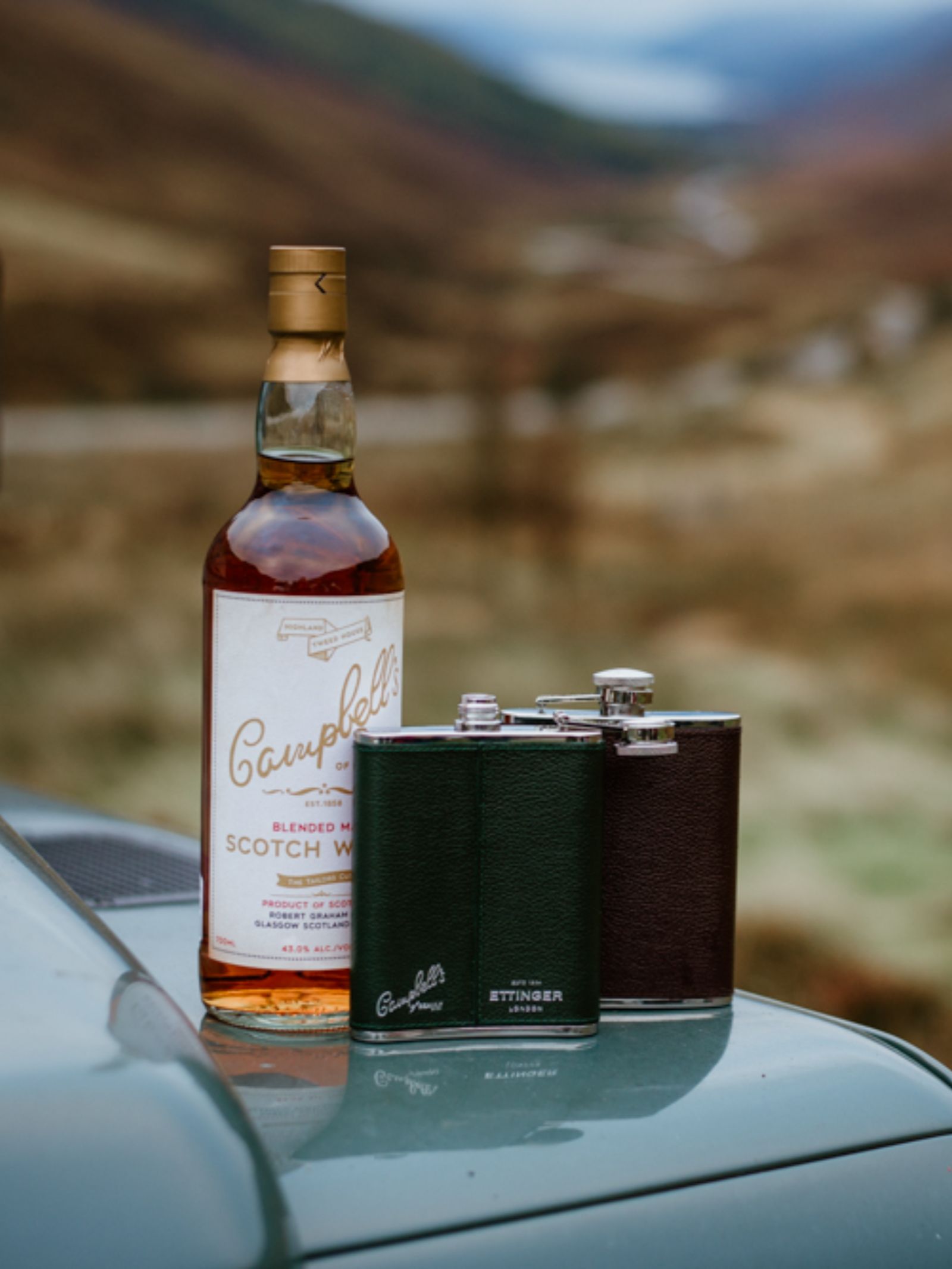 A bottle of Campbell’s blended Scotch whisky and two Campbell's of Beauly x Ettinger 6oz Forest Green Hip Flasks sit on a car hood, with a blurred scenic backdrop of hills and a winding road.