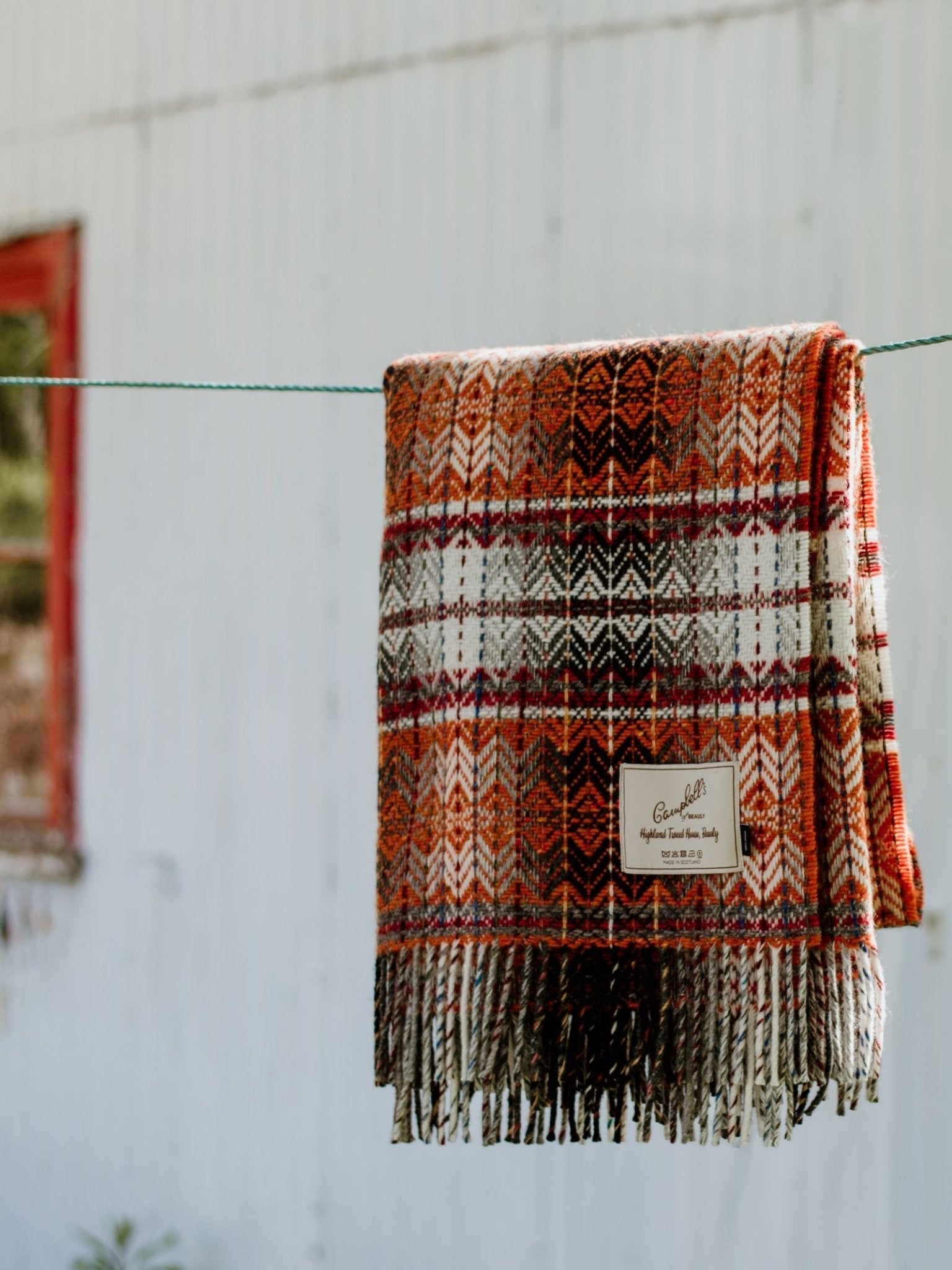 The Wyvis Lambswool Throw by Campbells of Beauly, with orange, brown, and white plaid, hangs on a clothesline outside against a white corrugated metal wall and a red-framed window.