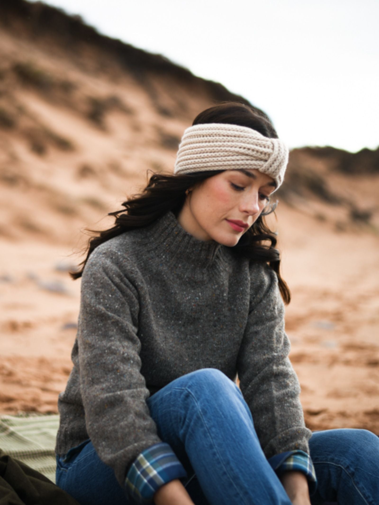 A woman in a gray sweater, blue jeans, and a Campbell's of Beauly Cashmere Ear Warmer sits on a sandy beach with dunes behind her—an ideal winter accessory for chilly days.