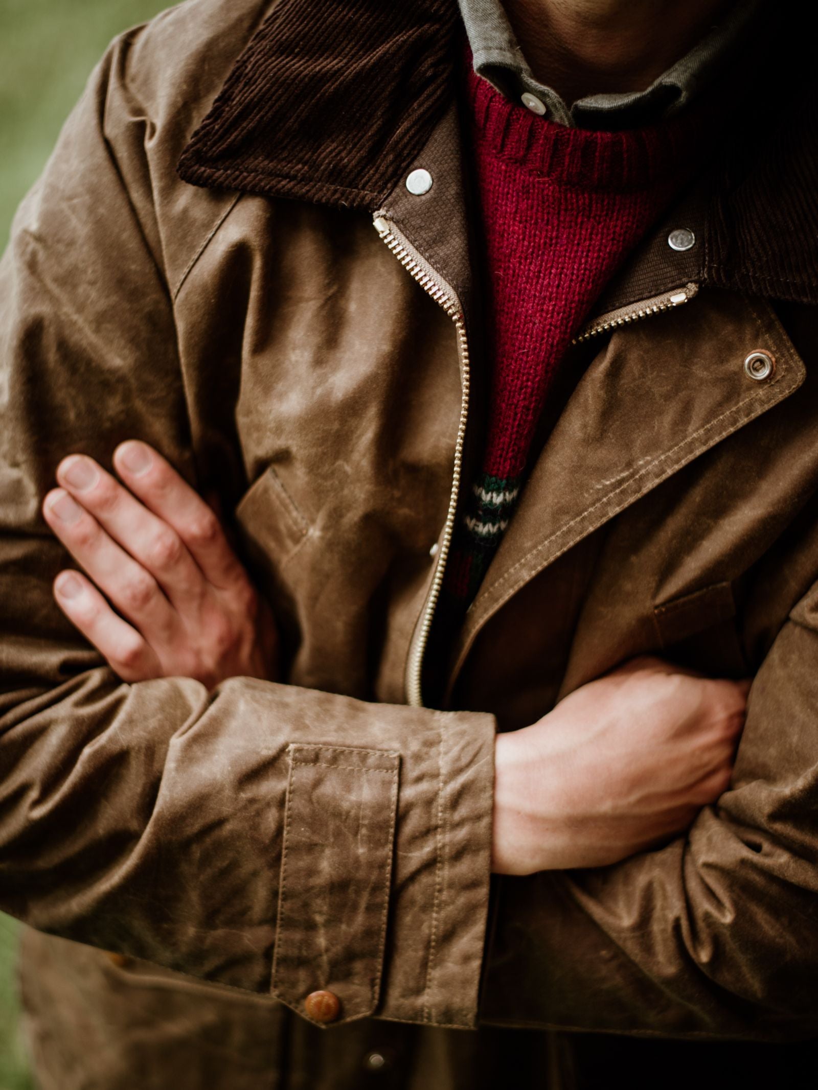 A person with arms crossed wears a Campbell's of Beauly Wax Jacket in brown waxed cotton, featuring a zipper and snap buttons, layered over a red knit sweater and collared shirt. They are outdoors with their face not visible.