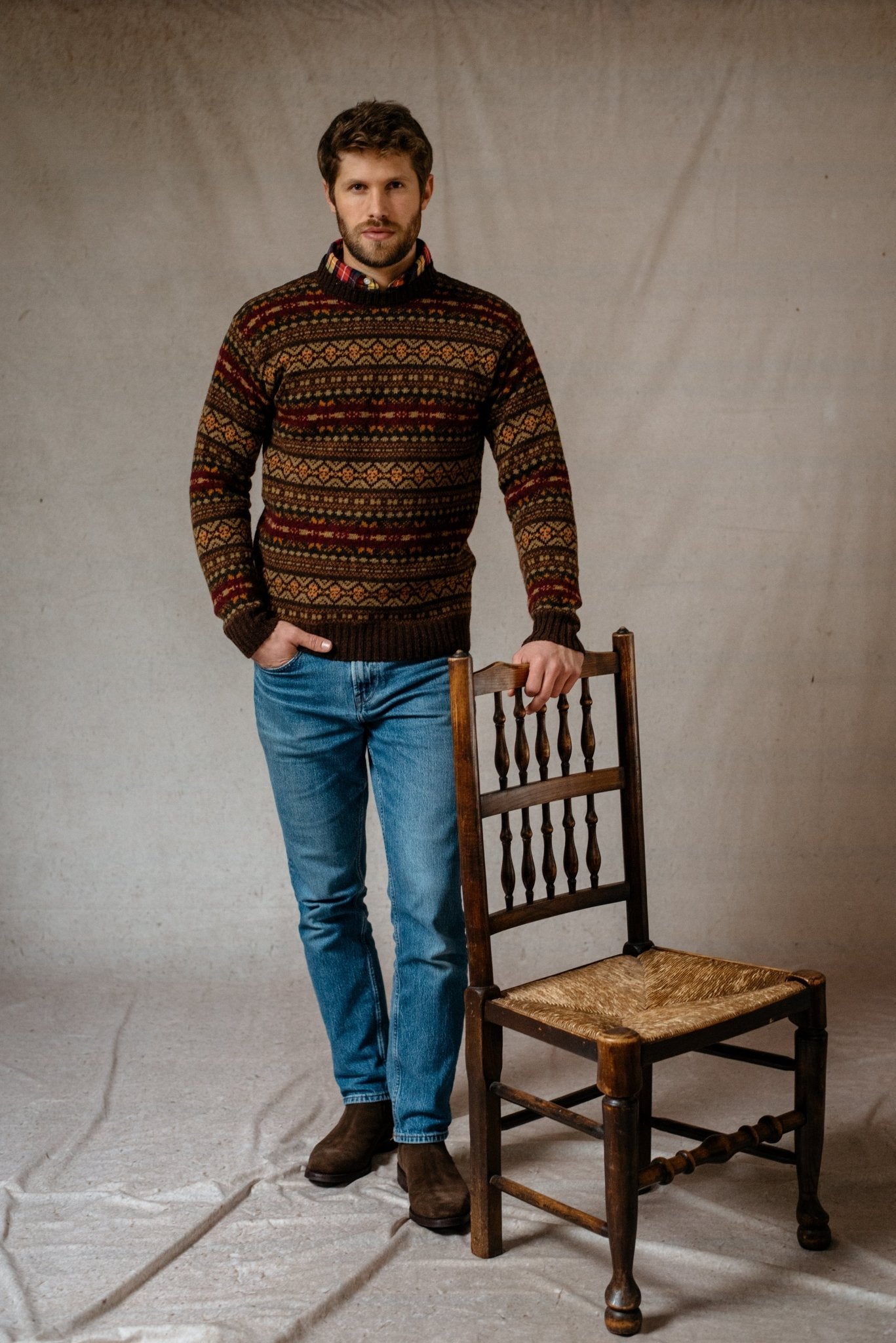 A man with a beard wears the Campbell's of Beauly Shetland Fairisle Crew Neck Jumper and blue jeans, standing by a wooden chair against a beige background. This classic knit adds stylish warmth to any winter look.