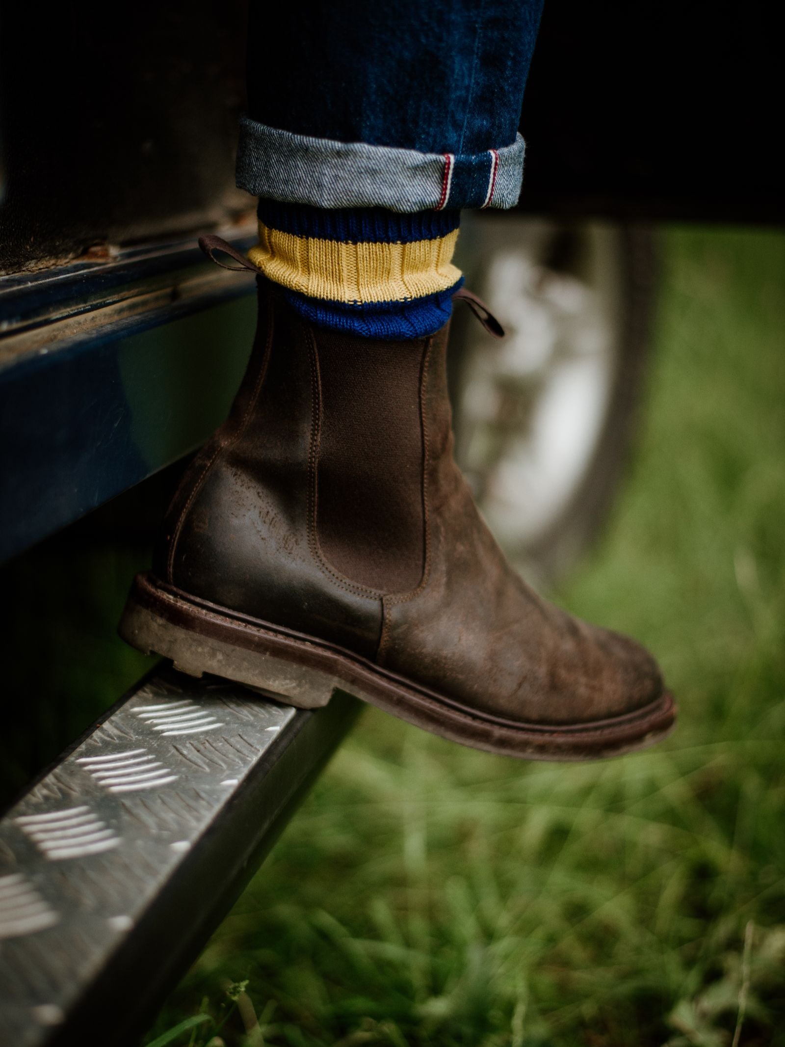 A person steps onto a metal footstep beside grass, wearing Campbell's of Beauly Lowlander Boots with Ridgeway soles and yellow socks with blue stripes. Denim jeans are rolled up above the ankle.