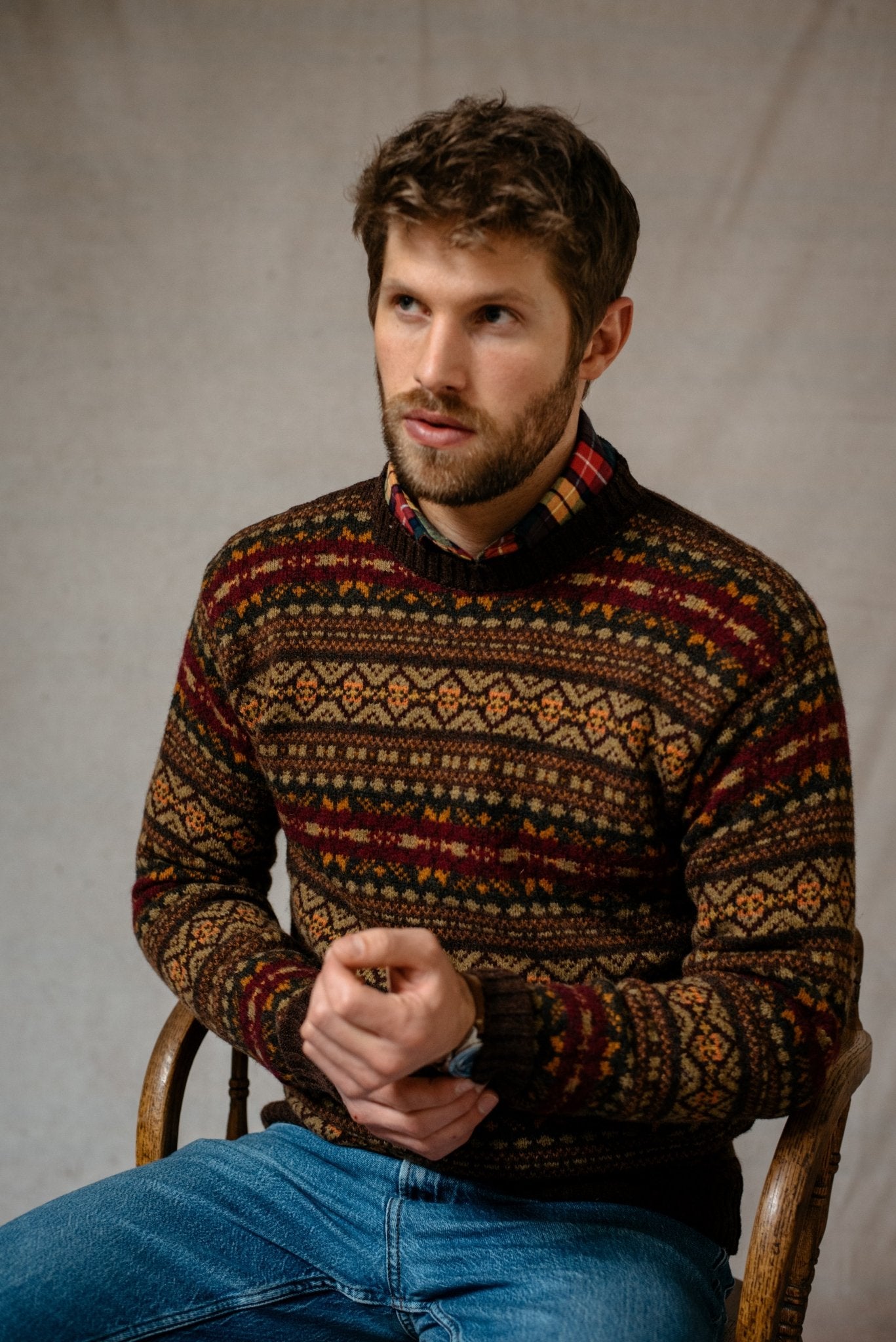 A man with short brown hair and a beard sits on a chair, wearing the Campbell's of Beauly Shetland Fairisle Crew Neck Jumper over a collared shirt and blue jeans. He looks slightly to the side, hands clasped, against a neutral background.