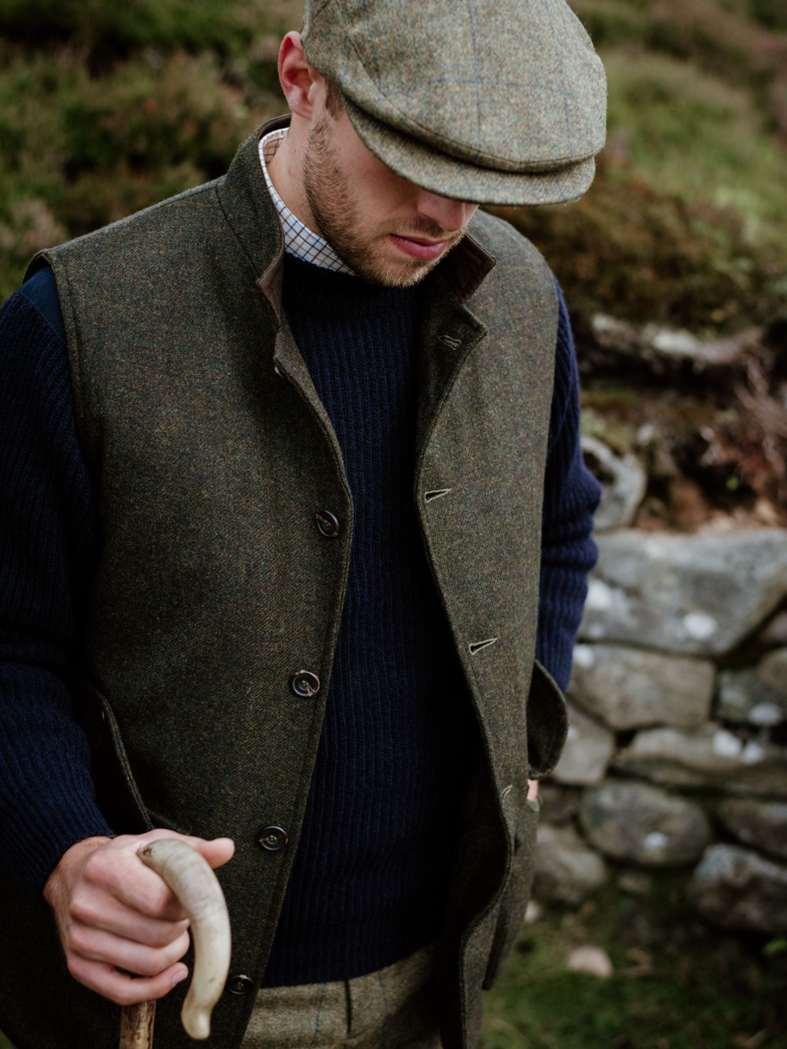 A man outdoors wears the Field Nehru Gilet by Campbells of Beauly over a navy sweater and checked shirt, pairing it with a tweed flat cap. He holds a wooden walking stick amid grass and a stone wall, evoking the rural countryside.