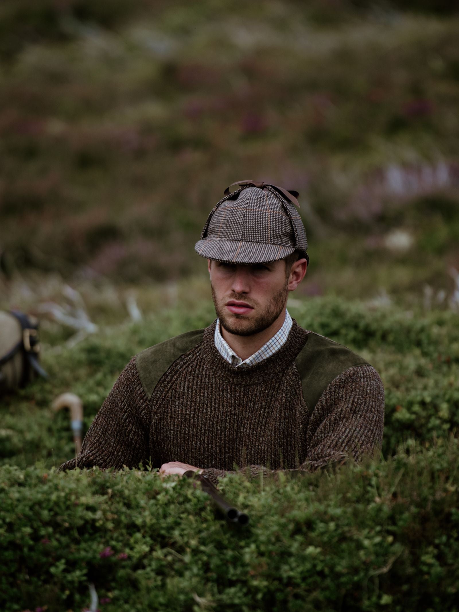A man in a Campbells of Beauly Sherlock Holmes Hat and brown knit sweater sits among green bushes, blending with the landscape. He looks contemplative as blurred grass and wildflowers frame him.