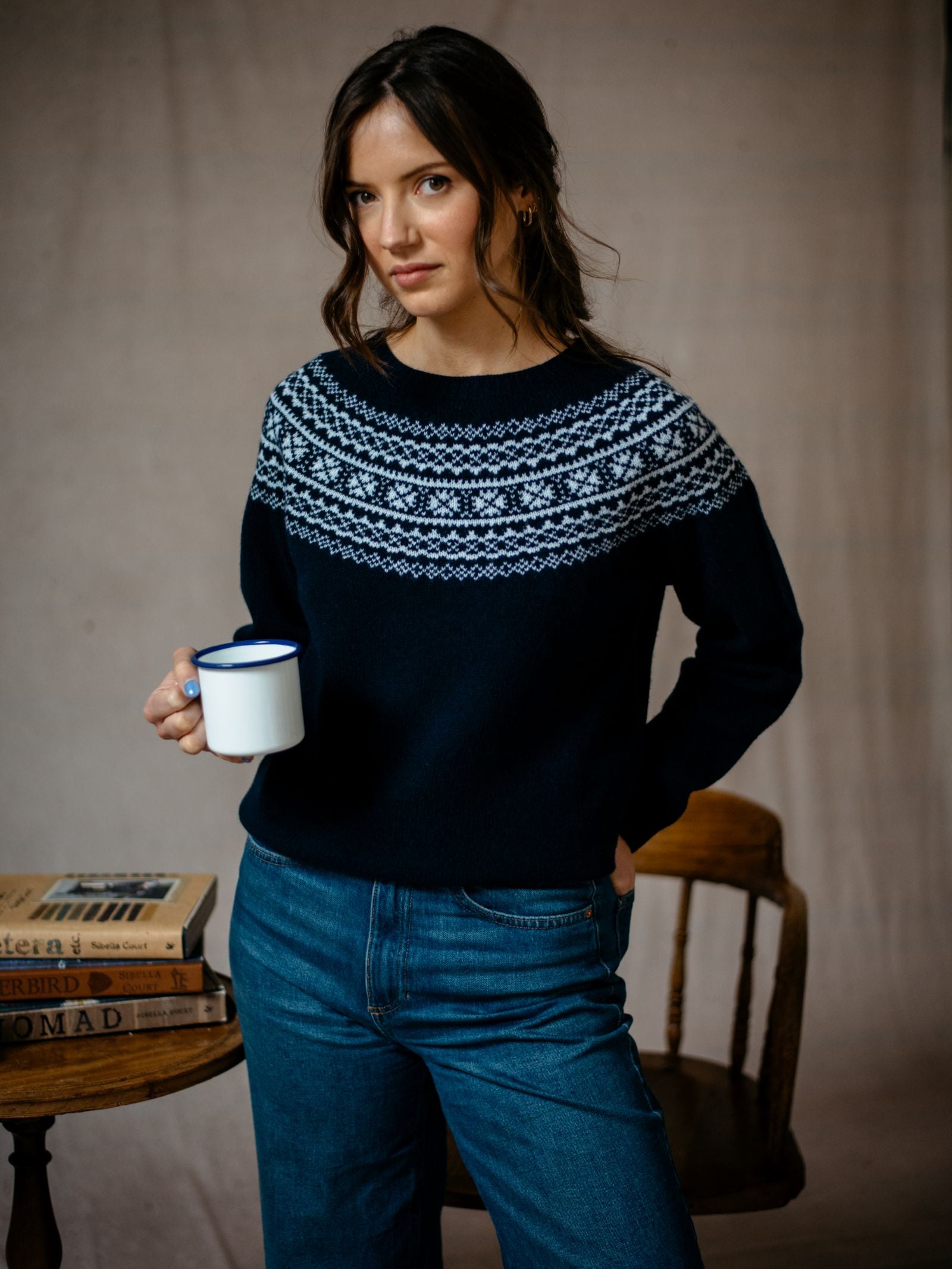 A woman with wavy brown hair wears Campbells of Beaulys Two-Colour Fairisle Yoke Jumper made from superfine Geelong lambswool. She stands indoors holding a white mug near a wooden chair and book-filled table against a softly lit backdrop.