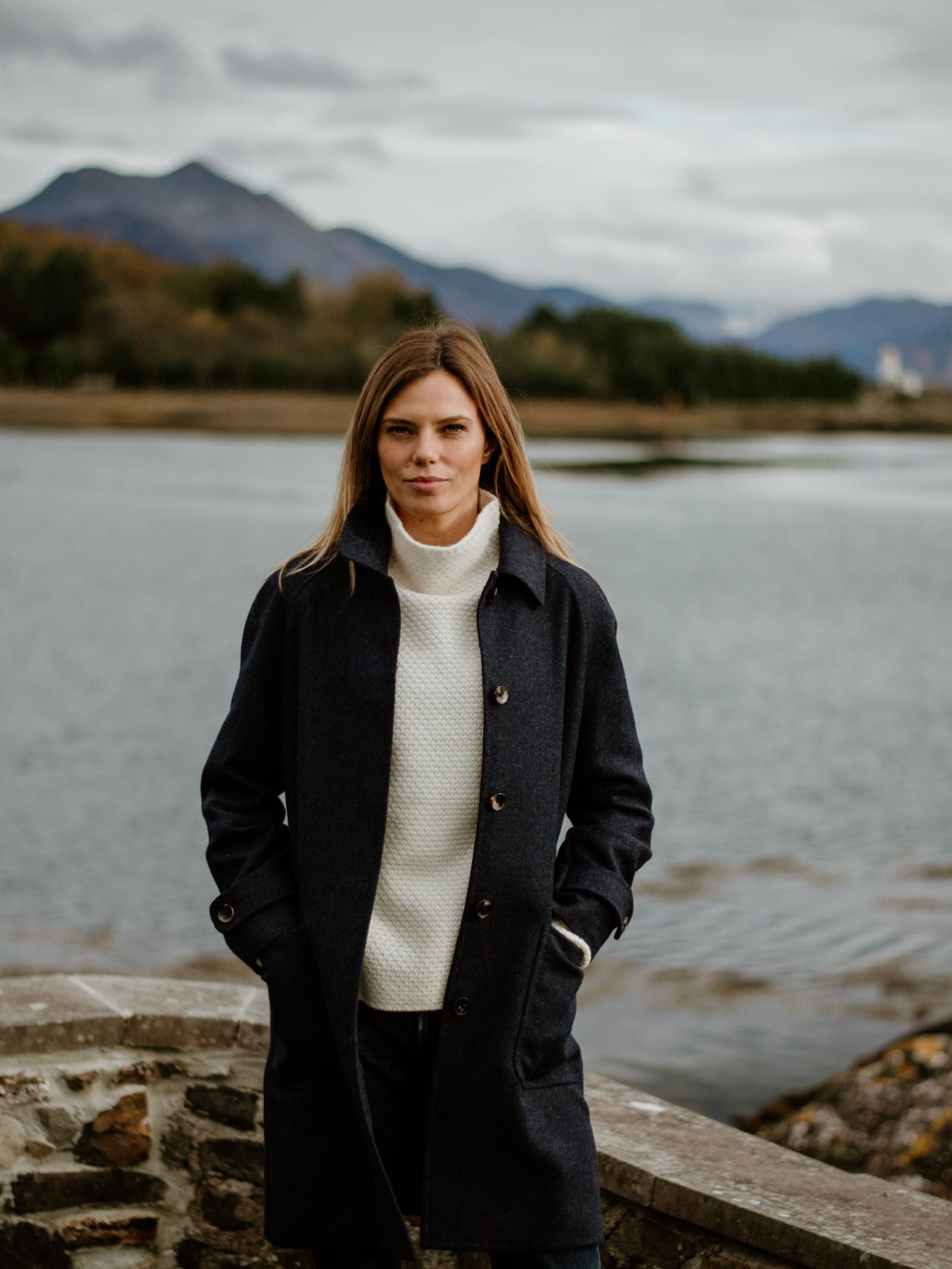 A woman with long brown hair stands outdoors by a stone wall and water, wearing the Campbells of Beauly Midnight Collar Coat over a white sweater. Mountains and trees are in the background under a cloudy sky.