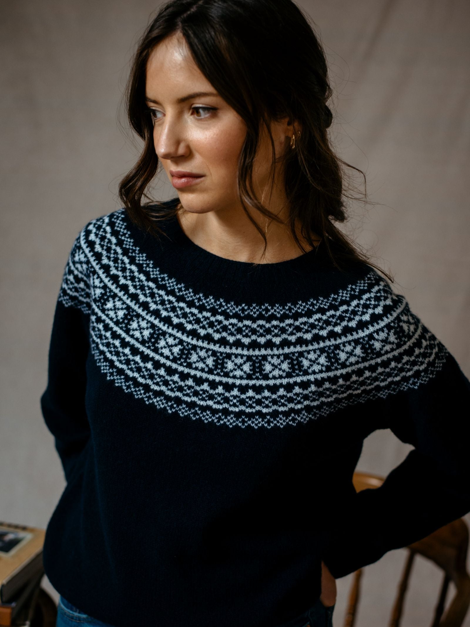 A woman with long brown hair styled in loose waves wears the Campbells of Beauly Two-Colour Fairisle Yoke Jumper, looking thoughtfully to the left against a softly lit neutral background.