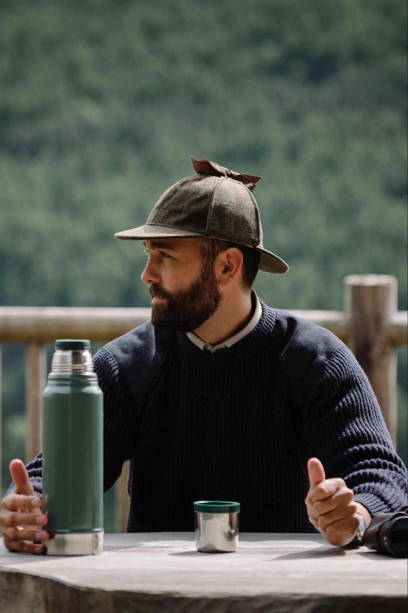 A man wearing a Campbells of Beauly Sherlock Holmes Hat and dark sweater sits at an outdoor table with a green thermos and metal cup, gazing to the side. A wooden railing and blurred forest complete the background.