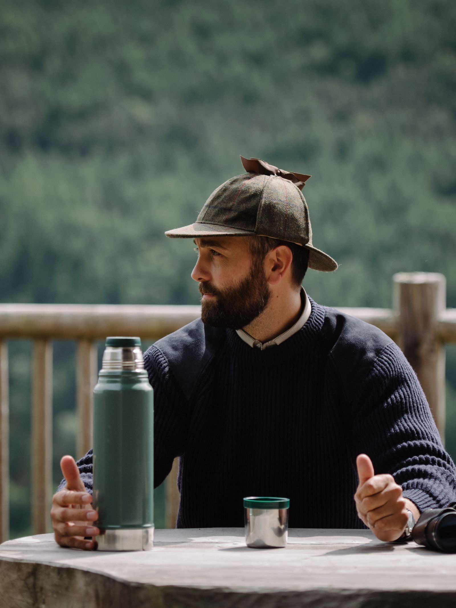 A bearded man in a knit sweater sits at a wooden table outdoors, wearing the Campbells of Beauly Sherlock Holmes Hat. With a green thermos and metal cup before him, forested hills in the background evoke heritage adventure.