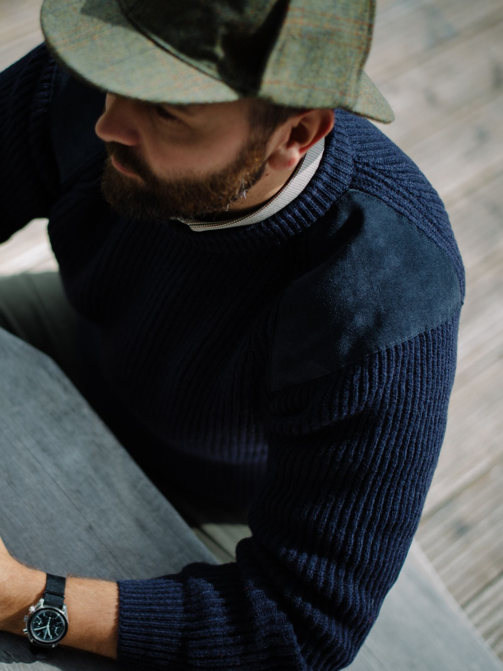 A man in a blue Campbells of Beauly Scotia Suede Jumper with suede shoulder patches sits at a gray table, wearing a greenish textured cap and black wristwatch. The photo is taken from above, showing wooden flooring in the background.