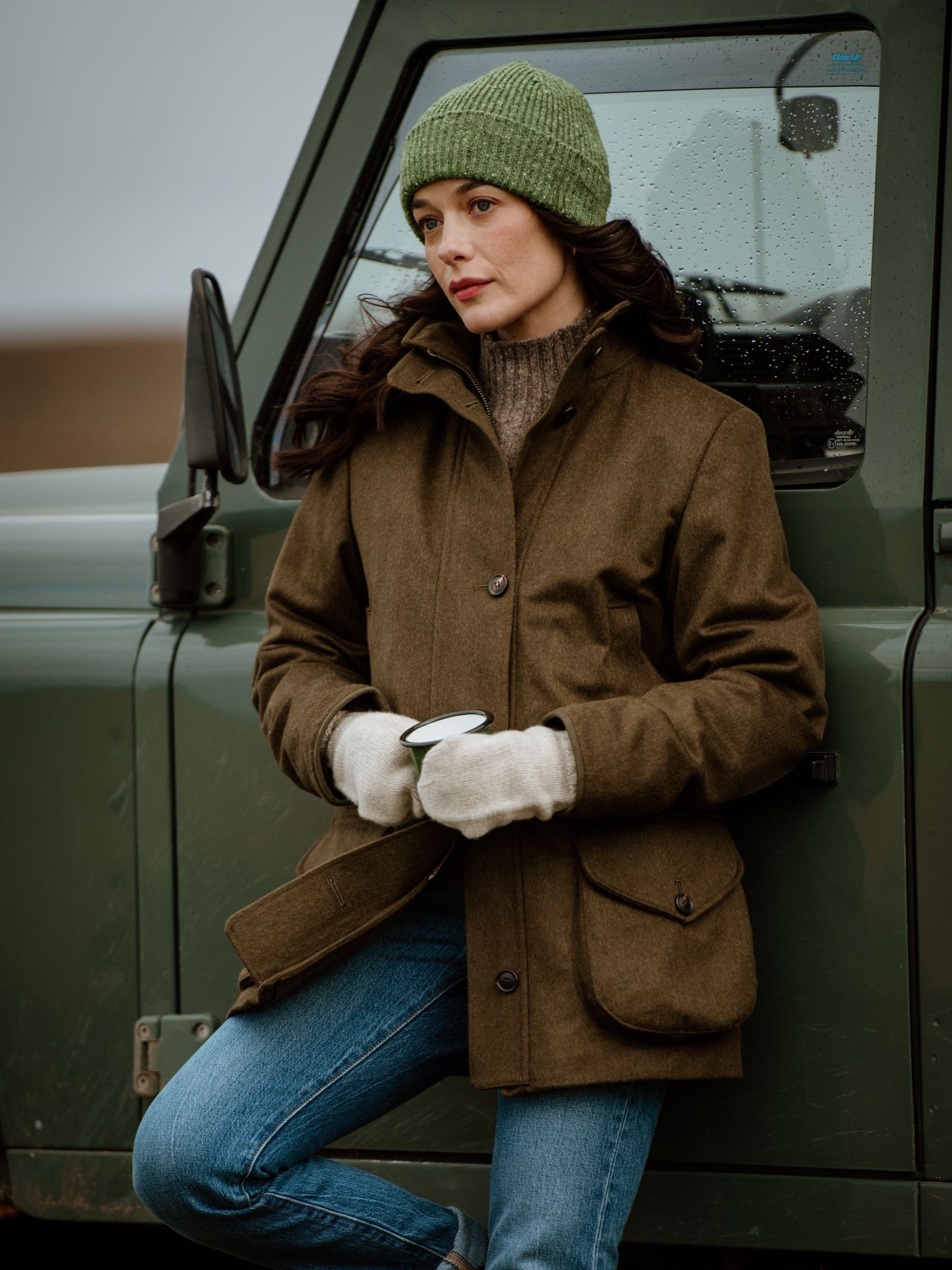 A woman wears a green Donegal Beanie by Campbell's of Beauly, paired with a brown jacket and gloves, leaning on a green vehicle with a mug. Her long brown hair flows over blue jeans as she enjoys a peaceful, cloudy day outdoors.