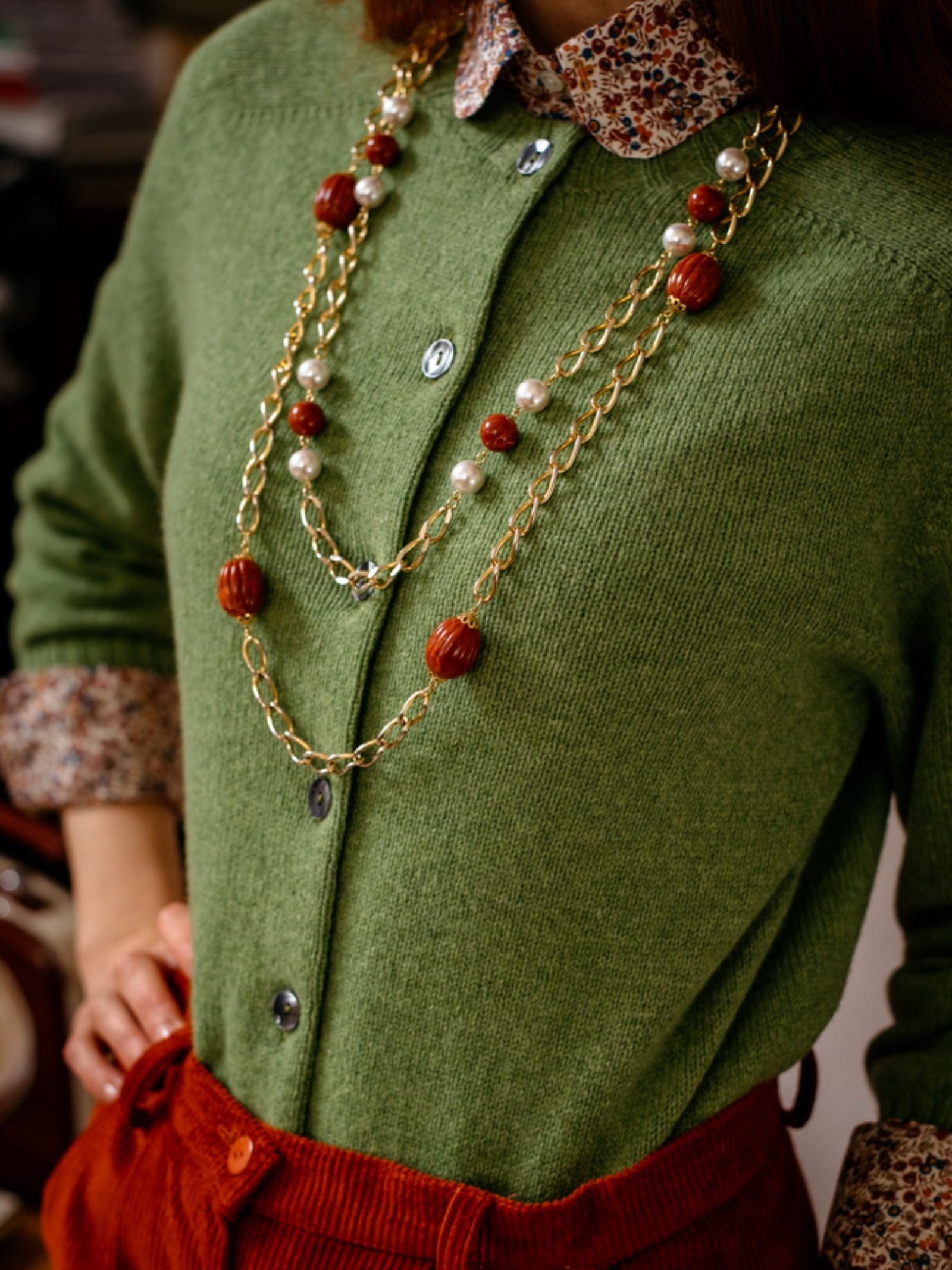 A person wears Campbells of Beauly Crew Cardigan in green over a floral-collared shirt, accessorized with layered gold and brown-beaded necklaces. High-waisted rust red corduroy pants are partially visible. The photo is cropped at the shoulders.