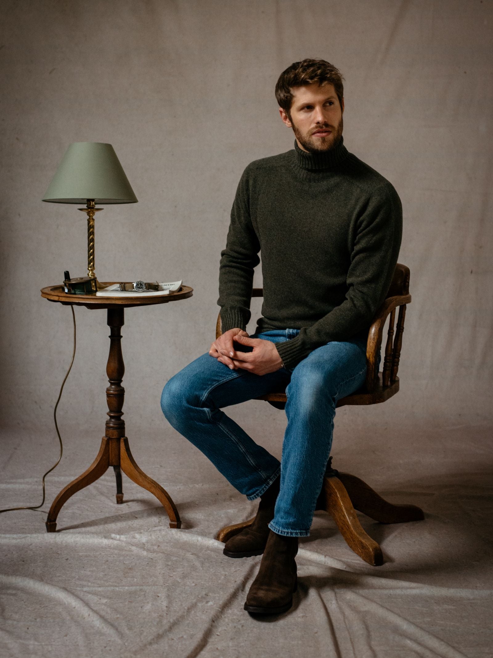 A man with short hair and a beard sits on a wooden chair, wearing the Campbells of Beauly Lambswool Poloneck in dark green, blue jeans, and brown boots. To his left is a small round table with a green lamp, glasses, and other small items.