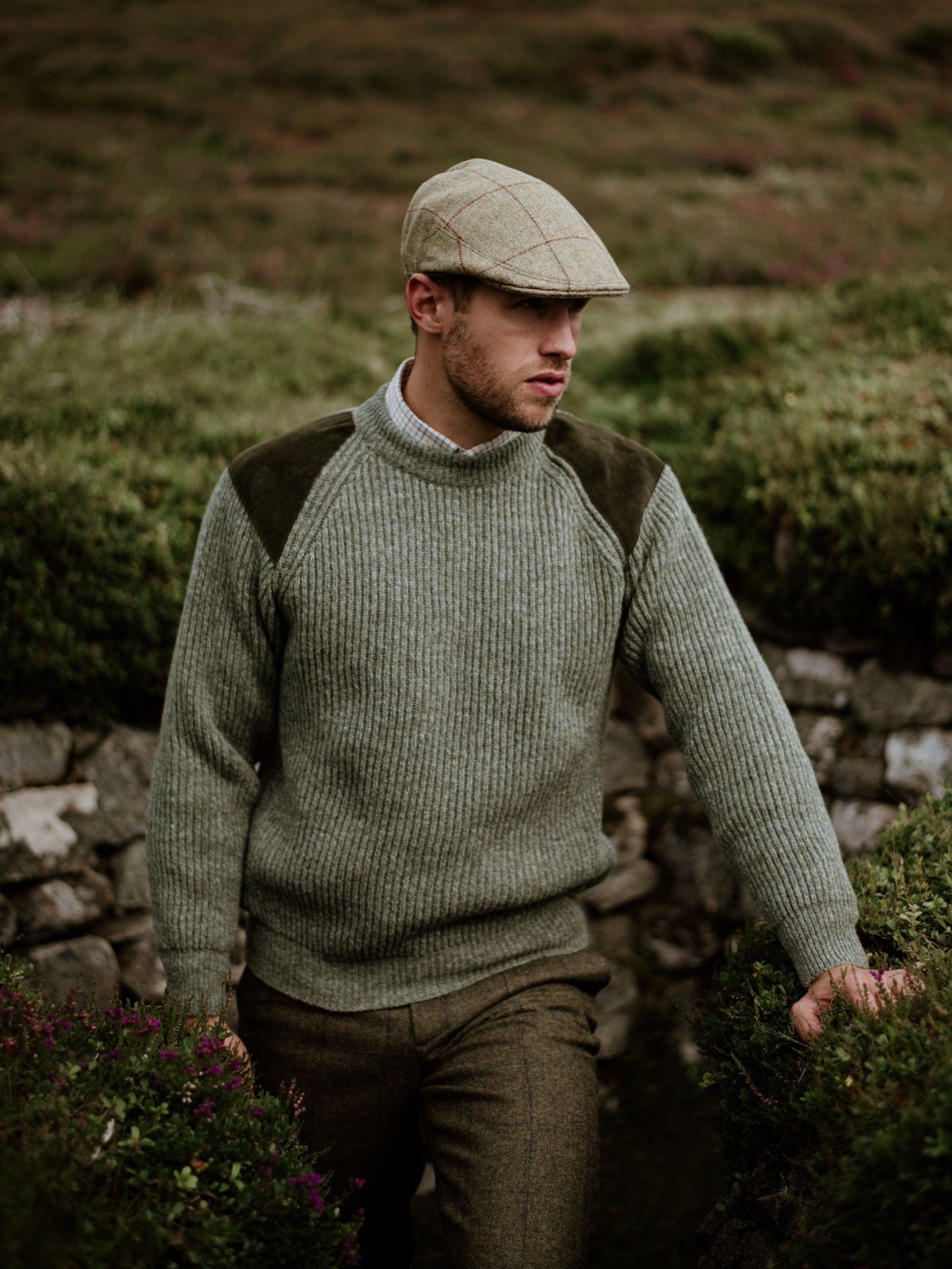 A man wearing Campbells of Beauly Scotia Suede Jumper with suede shoulder patches, a collared shirt, tweed trousers, and a plaid flat cap stands by green shrubs and stone walls, gazing thoughtfully in a rustic outdoor setting.