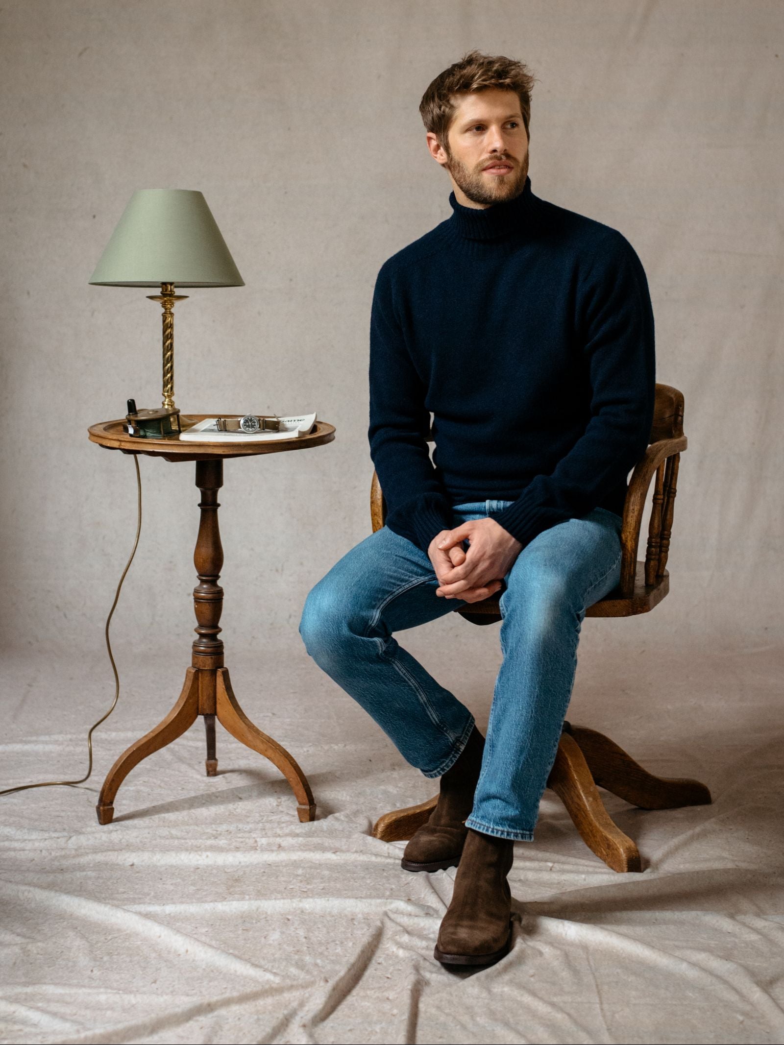 A man with light brown hair and a beard sits on a wooden chair, wearing the Campbells of Beauly Lambswool Poloneck, blue jeans, and brown boots. Next to him is a small wooden table with a green lamp, notebooks, and glasses against a neutral backdrop.