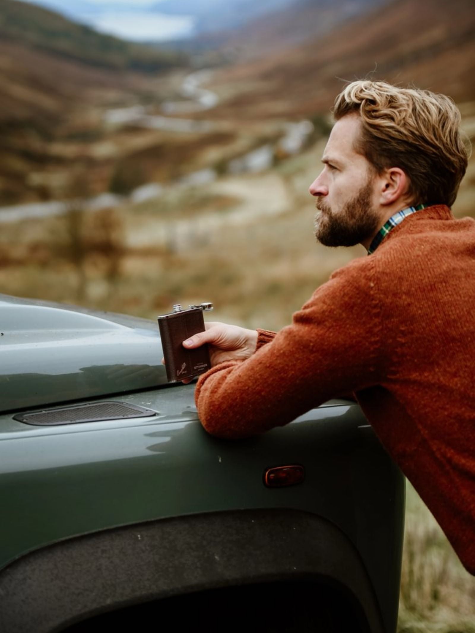 A man with light brown hair and a beard, in an orange sweater, leans on a green vehicle holding a Campbell's of Beauly x Ettinger 6oz Chocolate Hip Flask, gazing thoughtfully at the scenic mountains behind him.