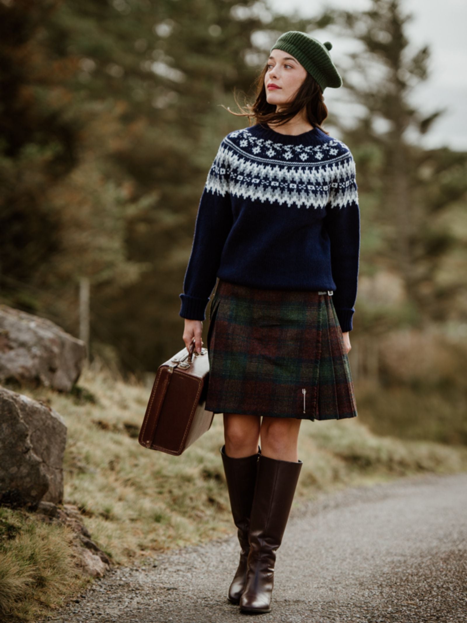 A woman wearing a Campbell's of Beauly Tweed Kilt, green beanie, navy patterned sweater, and tall brown boots walks along a country path with a brown suitcase, surrounded by trees and grass.