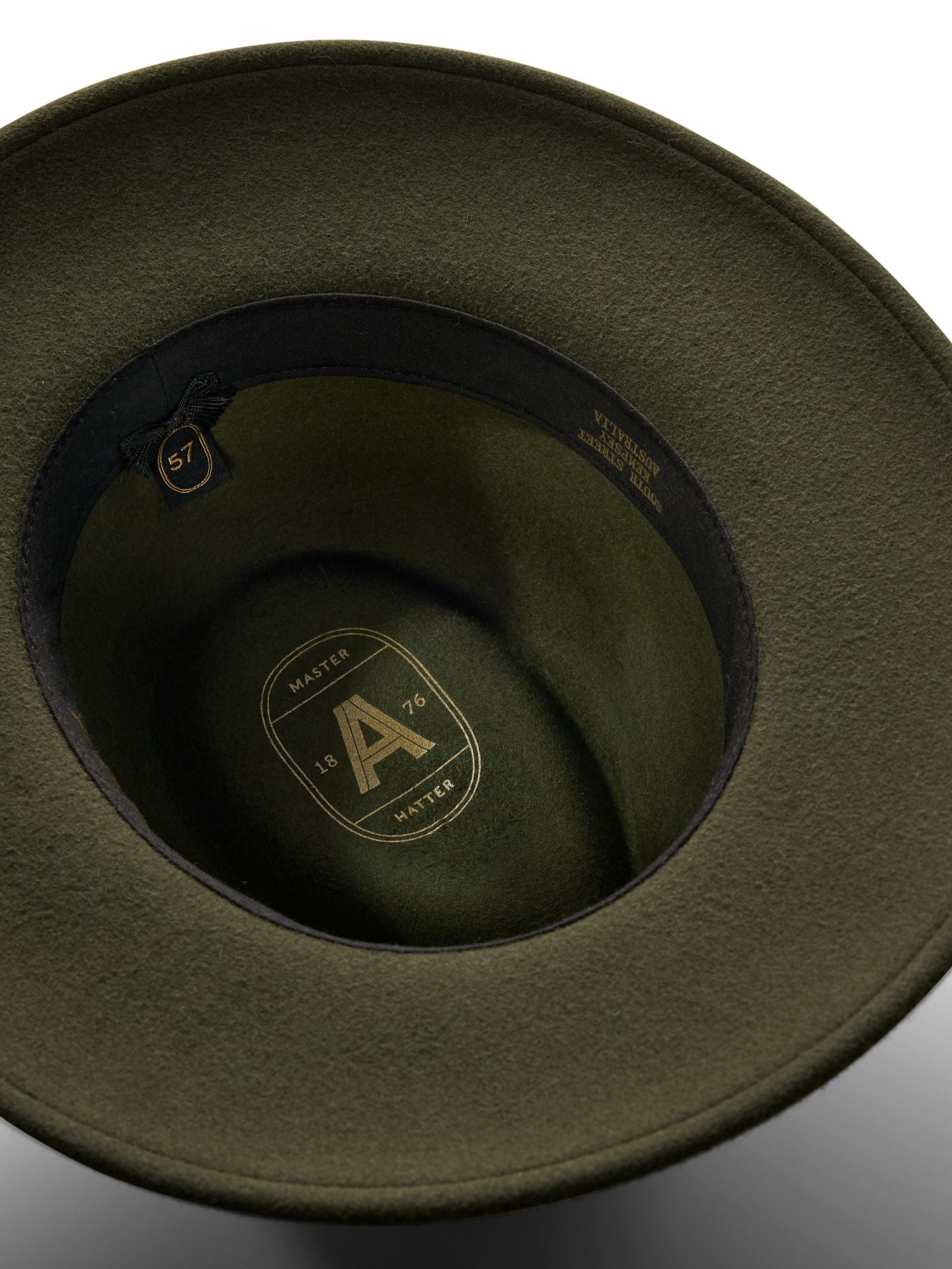 An overhead view of the Akubra Unisex Traveller Hat, made in Australia from olive green felt, showing its black inner band, gold 57 size tag, and gold lining with “A”, “1876”, and “HATTER” printed inside.