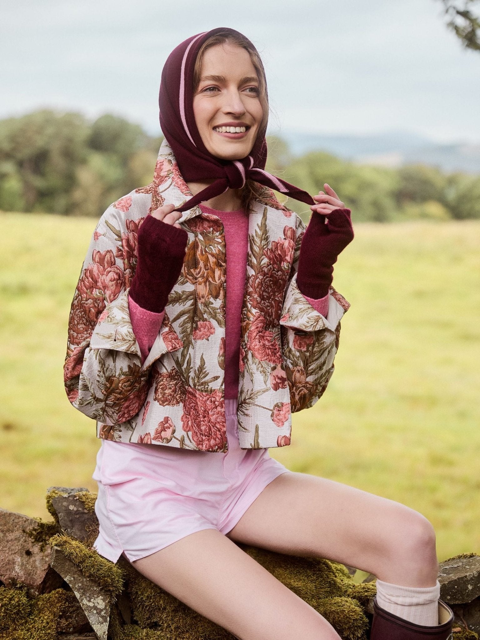 A smiling woman sits outdoors in green fields and trees, wearing a floral jacket, pink shirt, and shorts. She accessorizes with Campbell’s of Beauly Cashmere Neckerchief tied under her chin and matching gloves.