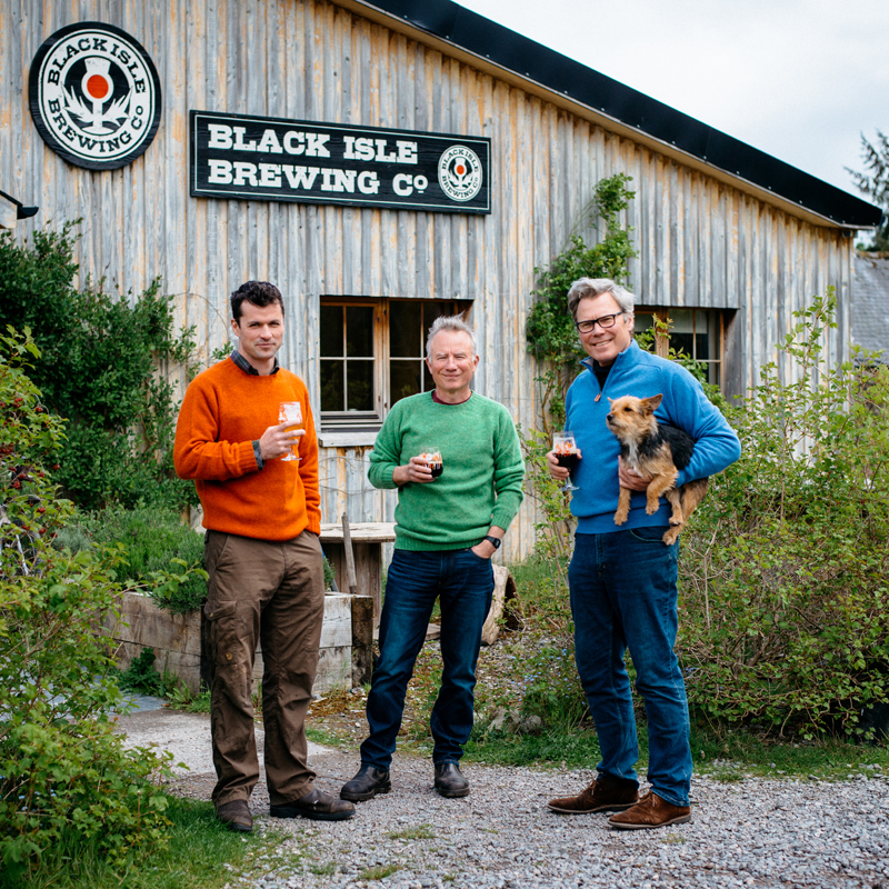 Three men stand outside Black Isle Brewing Co., each holding a drink. One man in a blue sweater holds a small dog. The wooden brewery building and greenery are visible in the background.