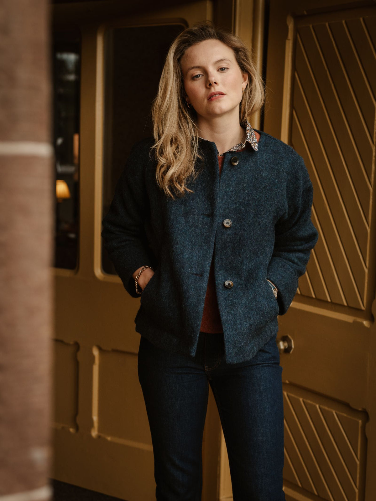 A woman with long blonde hair stands indoors, wearing the Campbell’s of Beauly Brushed Wool Jacket in navy and jeans, with her hands in her pockets, looking directly at the camera. Yellow paneled doors are in the background.