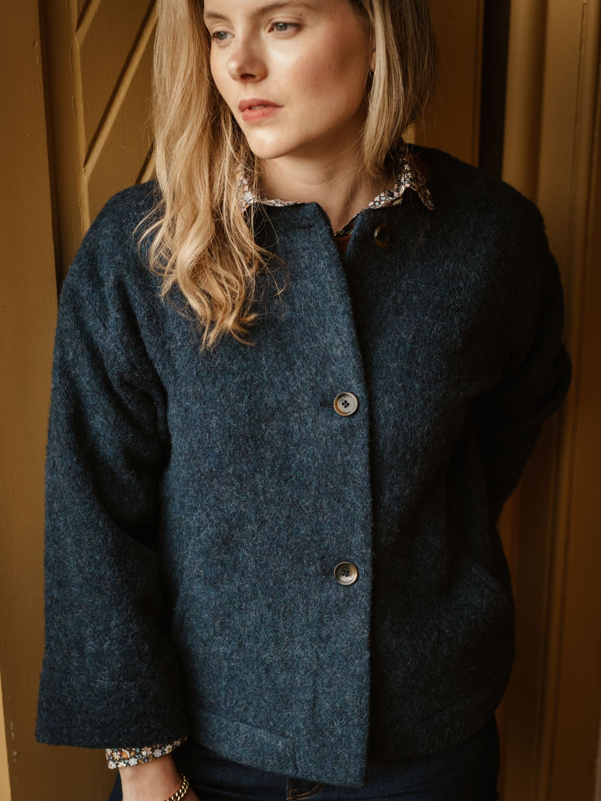 A woman with long blonde hair wears the Brushed Wool Jacket by Campbell’s of Beauly—a timeless, dark blue textured coat crafted from British boiled wool. She stands indoors against a backdrop of warm brown wooden panels, gazing to the side with a neutral expression.